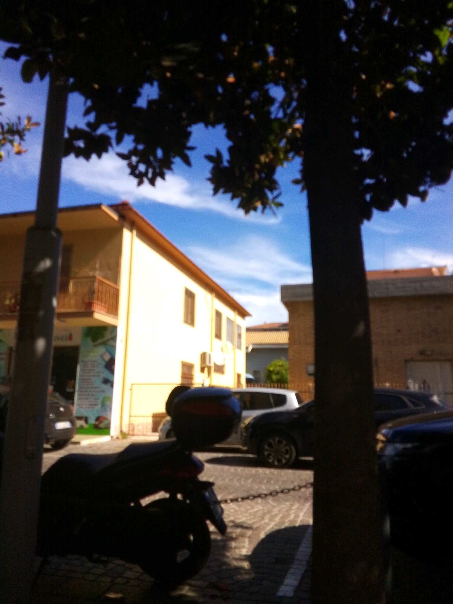 The image shows a street scene in Montesilvano, Italy, on a sunny day. In the foreground, a dark silhouette of a scooter with a top box is parked on a cobblestone street. To the left of the scooter, a light-colored pole, possibly a lamppost or utility pole, is visible. Behind the scooter, a light yellow building with a balcony and windows is present. Further back, another building, constructed of brick, is visible. The sky is blue with scattered white clouds. Trees with dark foliage cast shadows on the scene. Several cars are parked along the street, some of which are partially obscured by the scooter and the tree. The overall impression is a typical urban street in a Mediterranean climate.