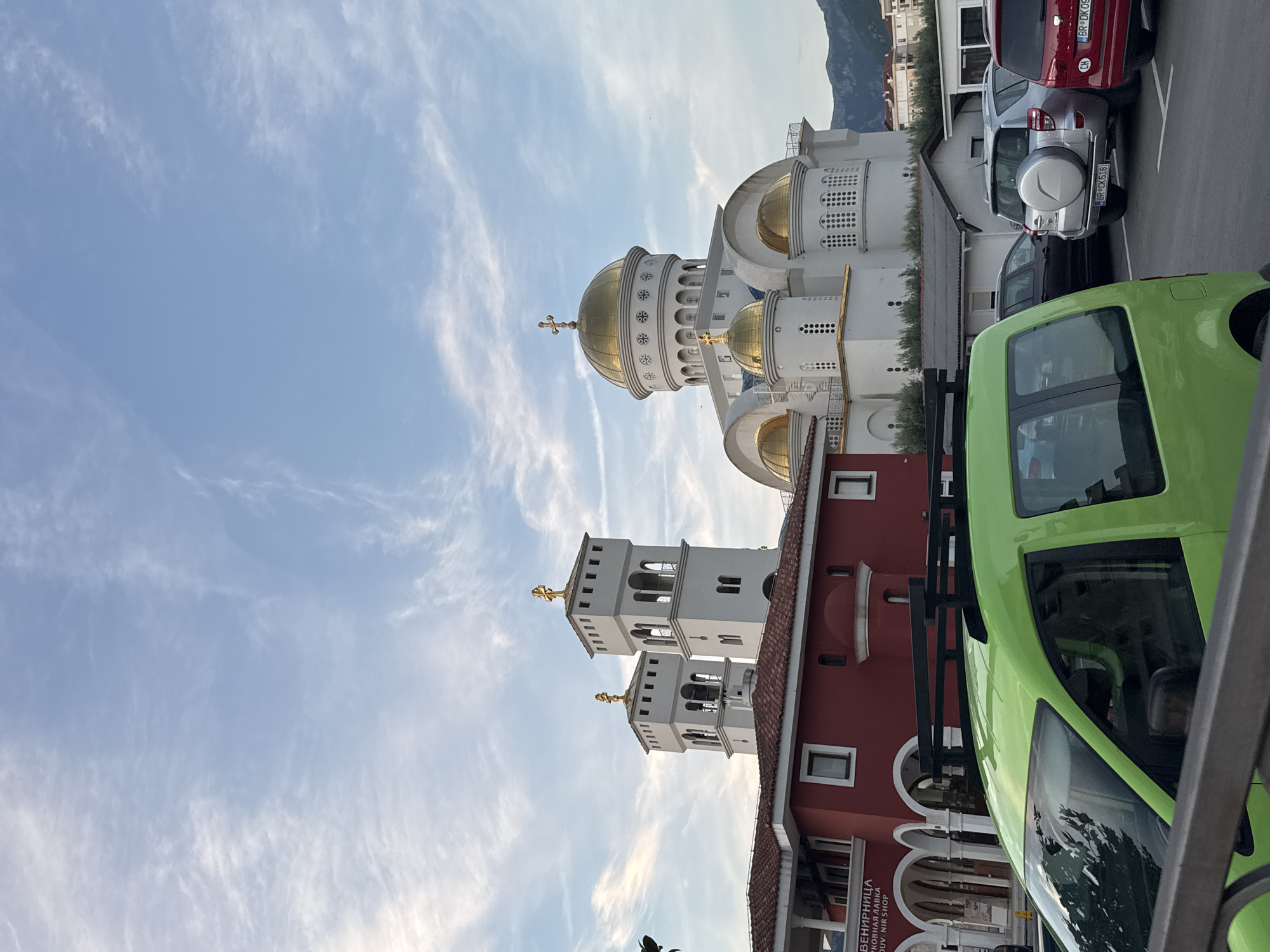 An upward-oriented outdoor shot captures a serene street scene in Šušanj, Montenegro, featuring a prominent Orthodox church complex under a partly cloudy sky. The main subject is a large, white church with three golden domes, each topped with a cross. The central dome is the largest and most intricately decorated, adorned with circular patterns and smaller crosses. To the left of the main church, two white bell towers with golden crosses at their peaks rise from a red-roofed building, possibly an adjacent chapel or a part of the complex.
In the foreground and mid-ground, several vehicles are parked on a paved surface. On the right, a bright lime green utility vehicle or small van is partially visible. Behind it, a silver Toyota RAV4 SUV is parked, displaying a Montenegrin license plate "BR DK518" (Bar region). Further back, a red hatchback, possibly a Renault Clio, also has a Montenegrin license plate "BR DK09" with a "CH" (Switzerland) sticker. Another dark-colored car is visible between the green vehicle and the silver SUV.
The sky is a clear blue with scattered, wispy white cirrus clouds, suggesting pleasant weather. The lighting indicates either late afternoon or early morning, casting a gentle glow on the buildings without harsh shadows. In the far background, faint outlines of mountains are visible, characteristic of the Montenegrin landscape. On the red-roofed building in the lower left, Cyrillic text is visible, reading "СУВЕНИРНИЦА ЦРКОВНА ЛАВКА", which translates to "SOUVENIR SHOP CHURCH SHOP", further confirming its association with the religious site. The overall atmosphere of the scene is calm and peaceful.