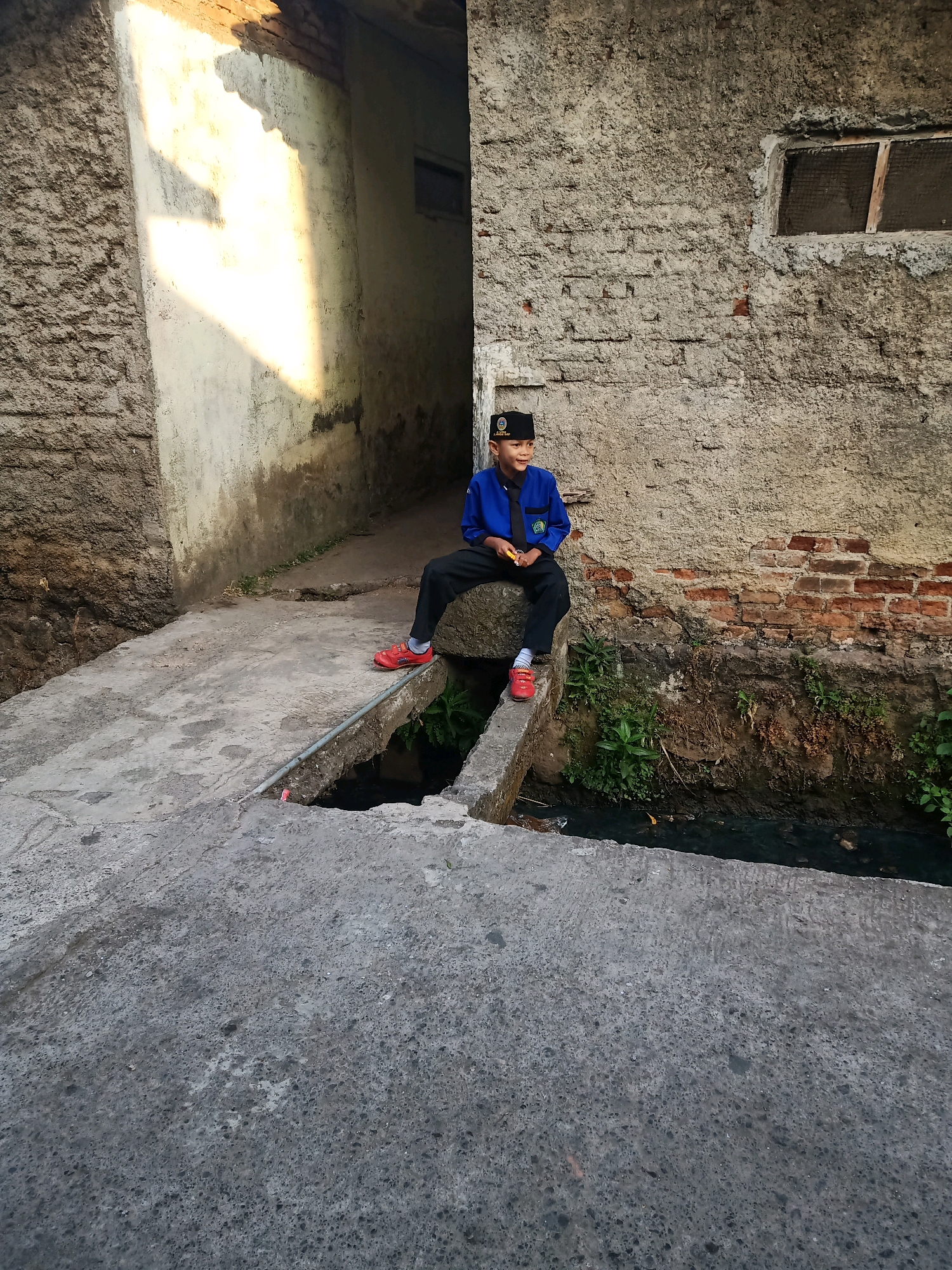 A young boy wearing a blue jacket, black pants, and a black peci (hat) is sitting on a concrete ledge. Sunlight streams in from the left, casting a strong shadow on a rough, textured wall behind him. The wall appears to be made of bricks and stucco, with a small, grimy window above and to the right of the boy. In front of the ledge, a dark, murky stream or ditch is visible with some green foliage growing along its edges. The ground in the foreground is also concrete, showing signs of wear and tear. The boy is looking off to his left with a slight smile. The scene appears to be in an alleyway or a narrow passage between buildings in an urban setting, likely in Garut, Indonesia, given the architectural style and the presence of the stream.