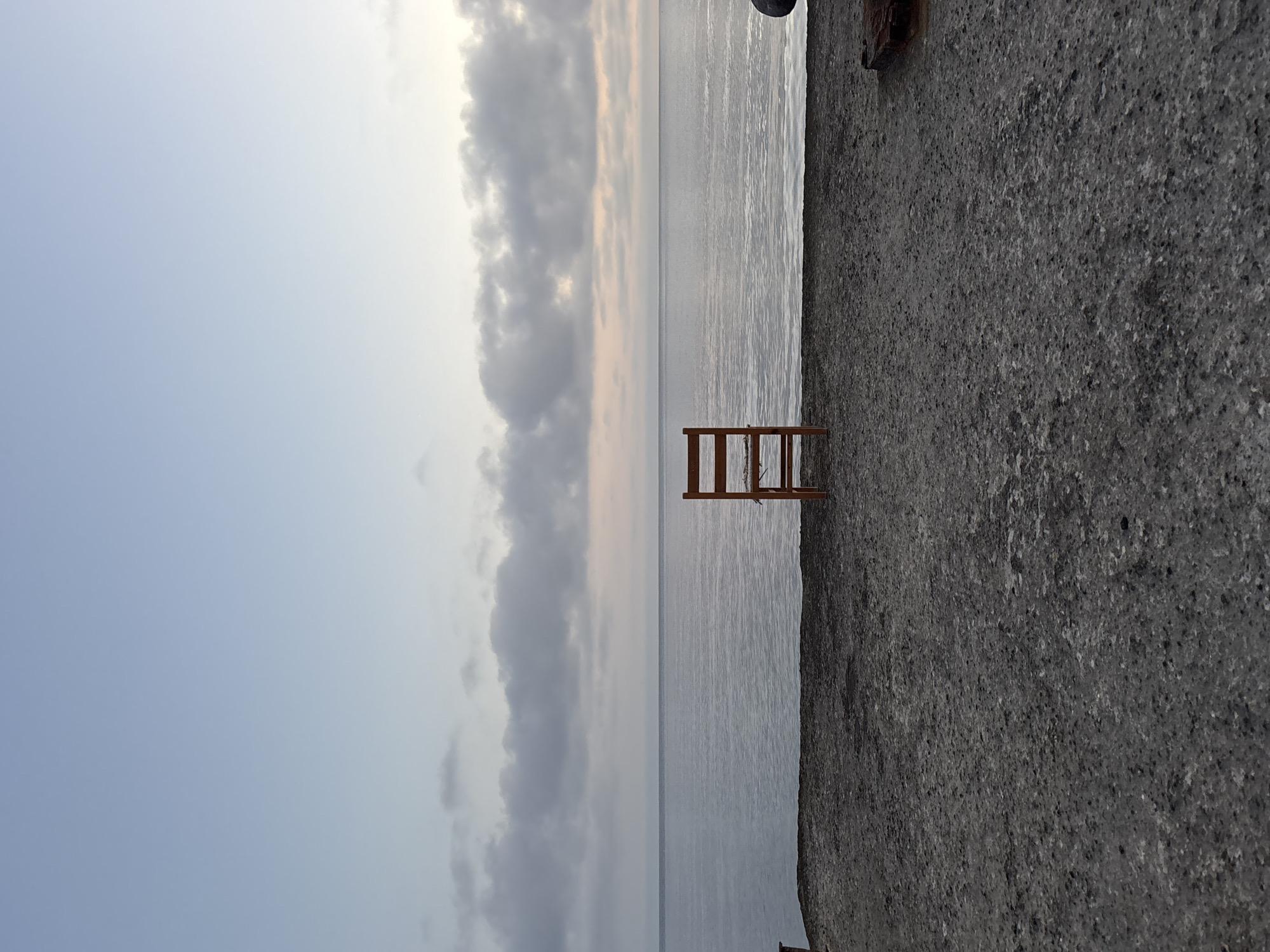 The image, oriented correctly, displays a serene coastal scene, likely in Lipari, Italy, characterized by soft, diffused lighting suggesting either dawn or dusk. The dominant feature is a simple wooden ladder-like structure positioned at the edge of a dark, coarse concrete or stone surface, leading down into calm, grey-toned water.
The upper portion of the frame is occupied by a cloudy sky. The clouds are a mix of light and darker grey, with hints of very soft pinkish-orange tones near the horizon line where the sky meets the water, indicating the early or late hour. Below the sky, a vast expanse of still, reflective water stretches across the midground. The water's surface shows subtle ripples, mirroring the muted light and clouds above.
The foreground consists of a roughly textured, dark grey surface, likely a concrete pier, seawall, or dock. The wooden structure, possibly part of a small pier or a set of steps for accessing the water, is centrally located on this surface and extends towards the water. No people are visible, and no specific activity is taking place, contributing to the quiet and contemplative atmosphere of the scene. There is no visible text in the image.