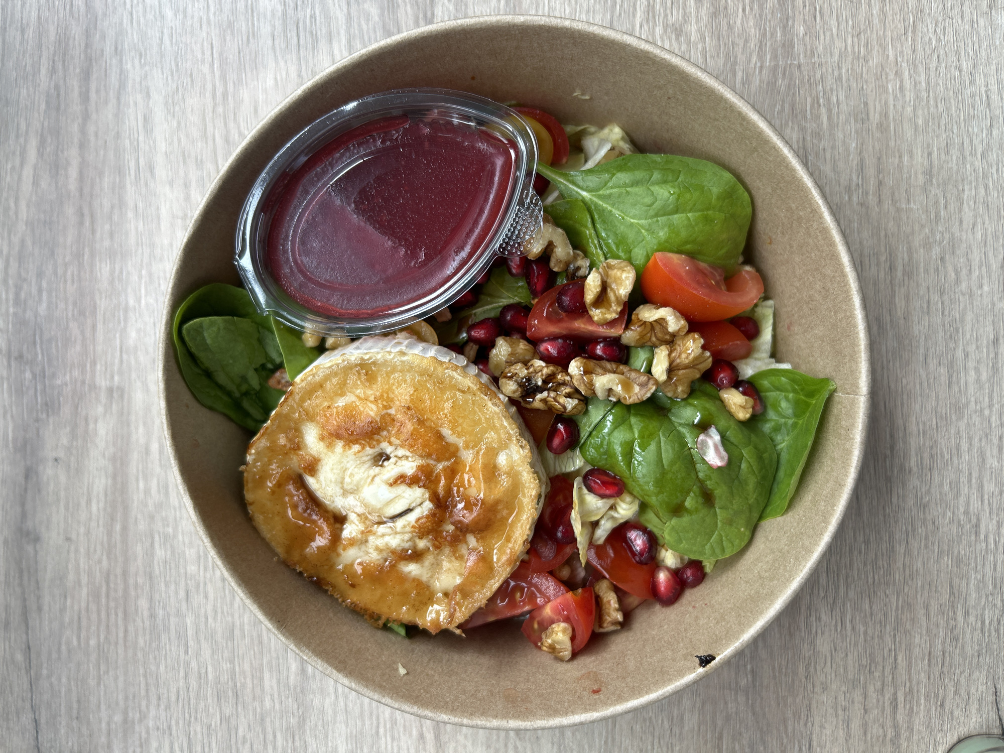 A top-down shot shows a salad in a cardboard bowl sitting on a light-colored, wood-grain surface. The salad includes a disk of baked or grilled cheese at the lower left. Other ingredients are spinach leaves, quartered tomatoes, walnuts, and pomegranate seeds. A small, clear plastic container of red dressing sits in the upper left of the bowl. The location is likely Sofia, Bulgaria.