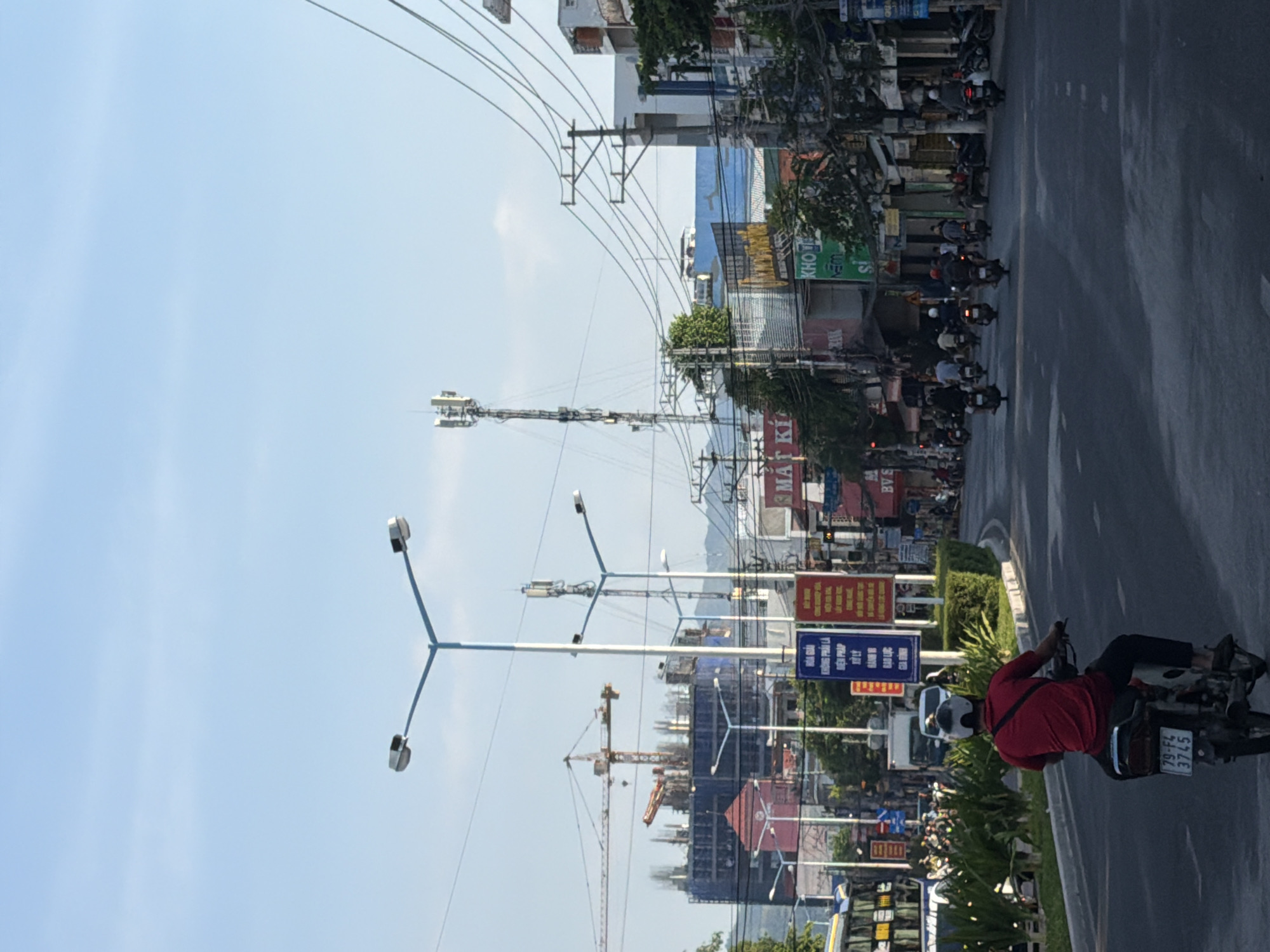 The image depicts a street scene in Bac Nha Trang Ward, Vietnam, under a clear blue sky. In the foreground, a person wearing a red shirt and dark pants rides a motorcycle with a license plate reading "79-F4 3745". The motorcycle is traveling away from the viewer on an asphalt road.
The road is lined with lush green plants and hedges on the left side. Beyond the vegetation, a bus is visible. The right side of the road features a row of buildings with various signs, some in Vietnamese. One sign prominently displays "MAT KI" and another appears to read "KHO". A large construction site with a crane and a partially built structure is visible in the background on the left.
Several other motorcycles and people are scattered along the street, suggesting moderate traffic. Tall telecommunication towers with antennas are present in the middle ground. Overhead, a complex network of electrical wires crisscrosses the sky. In the distance, a range of hills can be seen. The overall scene conveys a typical urban street environment in Vietnam, with a mix of modern infrastructure, ongoing construction, and daily life.