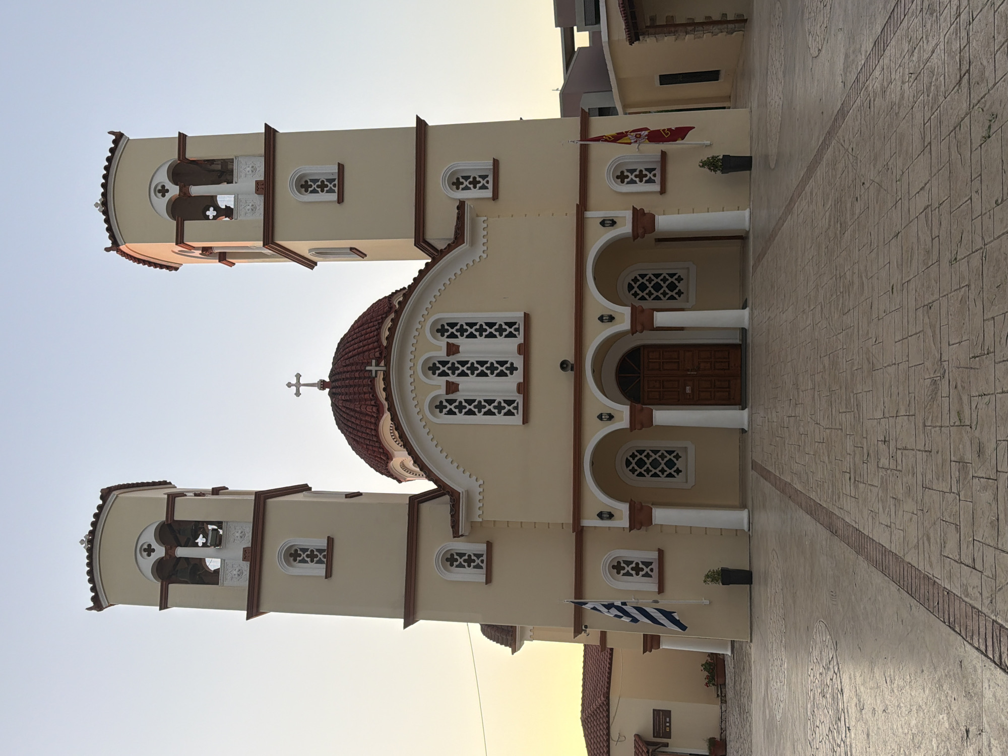 The image shows the Church of Saint George, a prominent landmark in Georgioupoli, Greece, bathed in the soft light of early morning or late afternoon. The church, painted a warm cream color with red-tiled roofs and dark red accents, stands majestically with two bell towers flanking a central dome topped by a cross. The bell towers feature open archways where bells are visible, and decorative cross-shaped windows. The façade of the church has multiple arched windows with intricate white latticework, and an arched entryway with double wooden doors. Two flags are displayed on the building: a Greek flag on the left and a red flag with an emblem on the right. The church is set within a paved courtyard, with patterned concrete slabs and brick borders. In the foreground, the paving has a circular pattern. The sky is a pale blue, transitioning to a soft yellow-orange on the left horizon, indicating the time of day. The environment appears serene and well-maintained, with potted plants adding touches of greenery. No people are visible in the image.