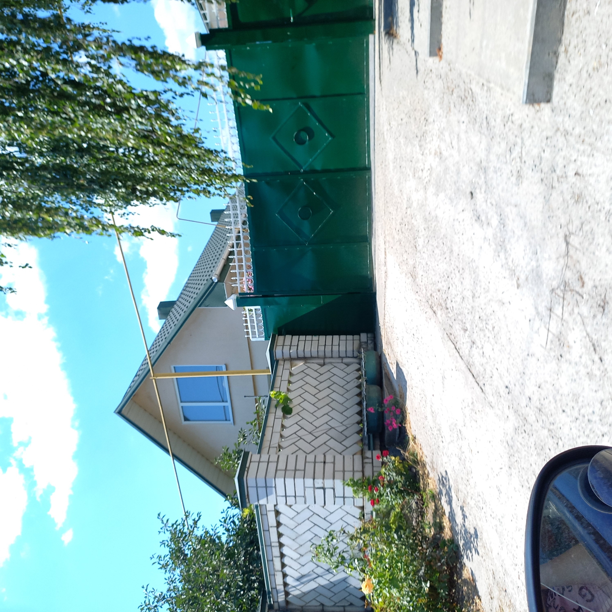 The image shows a street view in Klyntsi, Ukraine, on a bright, sunny day with a blue sky and scattered clouds. In the foreground, a driveway leads towards a property featuring a white brick fence with a patterned design. Adjacent to the fence are flower beds with red and yellow blooms. A large, dark green metal gate with a diamond-shaped embellishment stands to the right of the fence. Behind the gate and fence, a light-colored house with a green roof and a blue-paned window is partially visible. Trees, including one with hanging foliage, border the property and extend into the frame from the top right. The presence of a car's side mirror in the lower left corner suggests the image was taken from a vehicle.