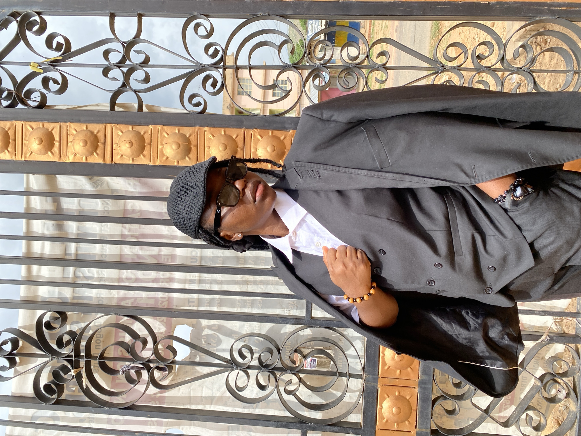 A person wearing a black suit, white shirt, black hat, and sunglasses is standing in front of an ornate metal gate. The person is adorned with bracelets on their wrist. The gate has golden-colored square patterns at the bottom and a complex scrollwork design in the middle and top sections. There is a newspaper behind the metal gate. In the background, there are buildings and visible sky. The location is Gwong, Nigeria.