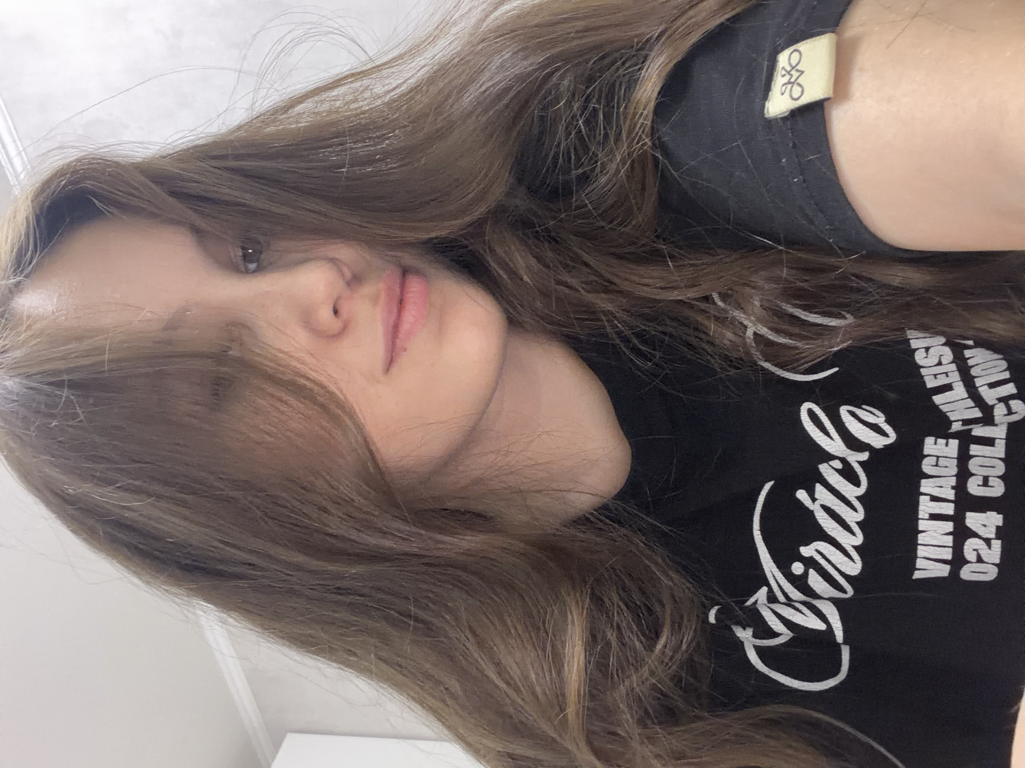 A medium shot is captured from a slightly high angle, focusing on a young woman with long, light brown hair. Her hair is styled loosely, with strands falling over her face and shoulders. She is wearing a black t-shirt with white lettering that appears to say "Miracle," "Vintage," and "Collection" across the chest. There is also a small, light-colored tag with a symbol on the sleeve. The woman's face is partially obscured by her hair, but her right eye is visible, and her lips are slightly parted. The background is a plain, light-colored wall with some subtle texture, suggesting an indoor setting. The overall lighting is soft and diffused. No specific location in Naberezhnye Chelny, Russia, can be identified from the image.