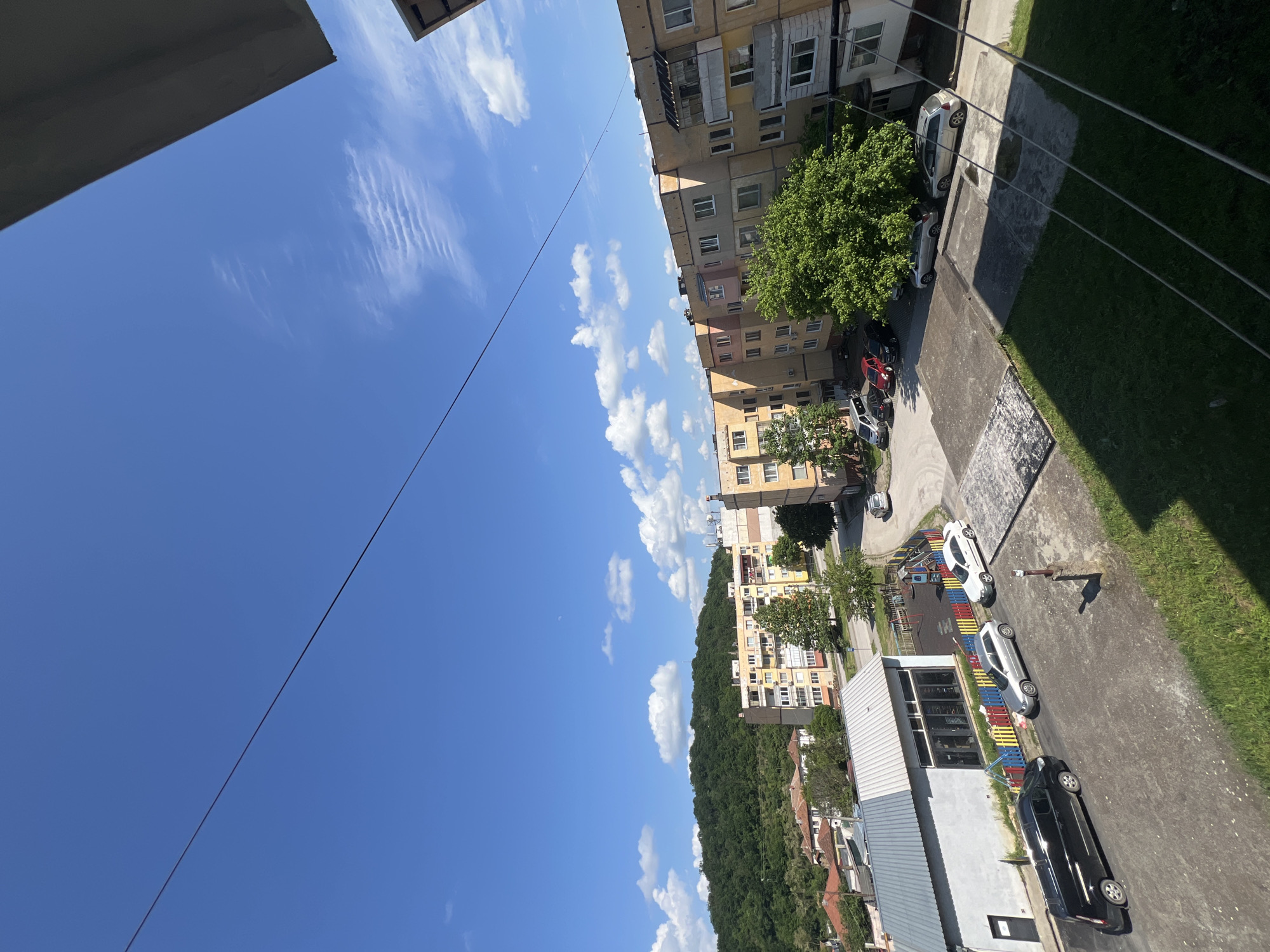 The image depicts an urban scene in daylight, likely from an elevated vantage point, such as a balcony. The sky is blue with scattered white clouds. An overhead wire extends diagonally across the frame. Several multi-story residential buildings, varying in color from yellow to orange, are visible. The buildings appear to be in a residential area, with trees interspersed. Cars are parked along the road. A playground with colorful structures is also visible. Beyond the buildings, a densely wooded area extends towards the horizon. The photo is taken in the city of Roman, Bulgaria.
