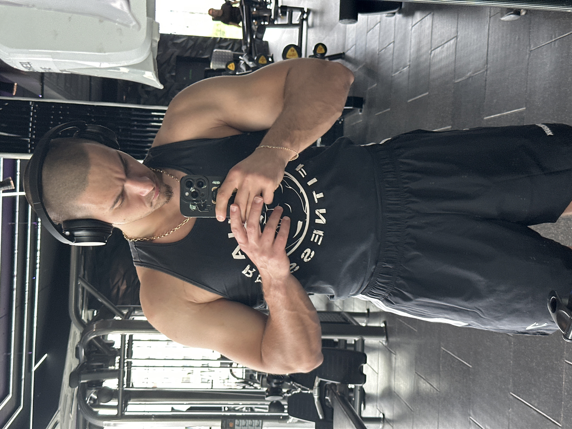 A man takes a selfie in front of a mirror in a gym, likely in Sofia, Bulgaria. He is wearing a black tank top, black shorts, and headphones, and has a gold chain. The gym has weight machines and gray flooring. The reflection in the mirror shows gym equipment.
