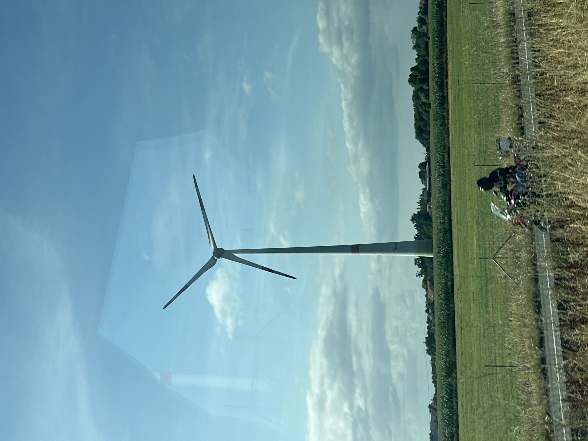 A vertical, slightly angled shot captures a tall wind turbine with three blades rotating against a blue sky dotted with scattered white clouds. The turbine is white with red bands near the top and bottom of its shaft, and some faint text is visible on the lower part of the shaft. In the foreground, a green grassy field stretches towards a line of trees and buildings in the distance. A motorcycle with a rider wearing a black helmet is visible on a road running through the field. The image is taken through a reflective surface, possibly a window, as indicated by faint reflections in the sky. The scene appears to be a rural landscape. The location is identified as Wuustwezel, Belgium.