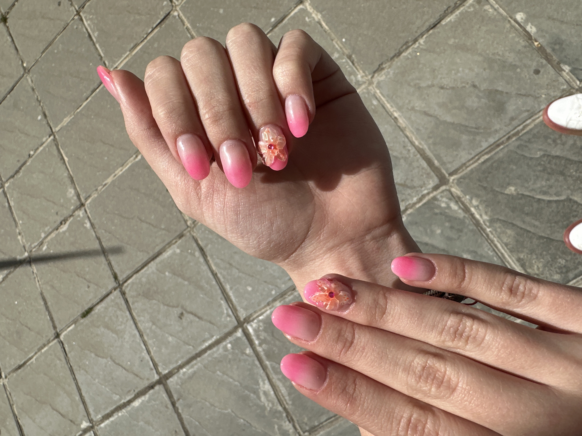 A person's hands are displayed in the image, showcasing pink ombre-style manicured nails. On one of the fingers, there is a raised floral design. The hands are positioned over a gray, tiled outdoor surface, possibly a sidewalk or patio. The scene is lit by sunlight. The exact location in Sofia, Bulgaria, cannot be determined from the image alone.