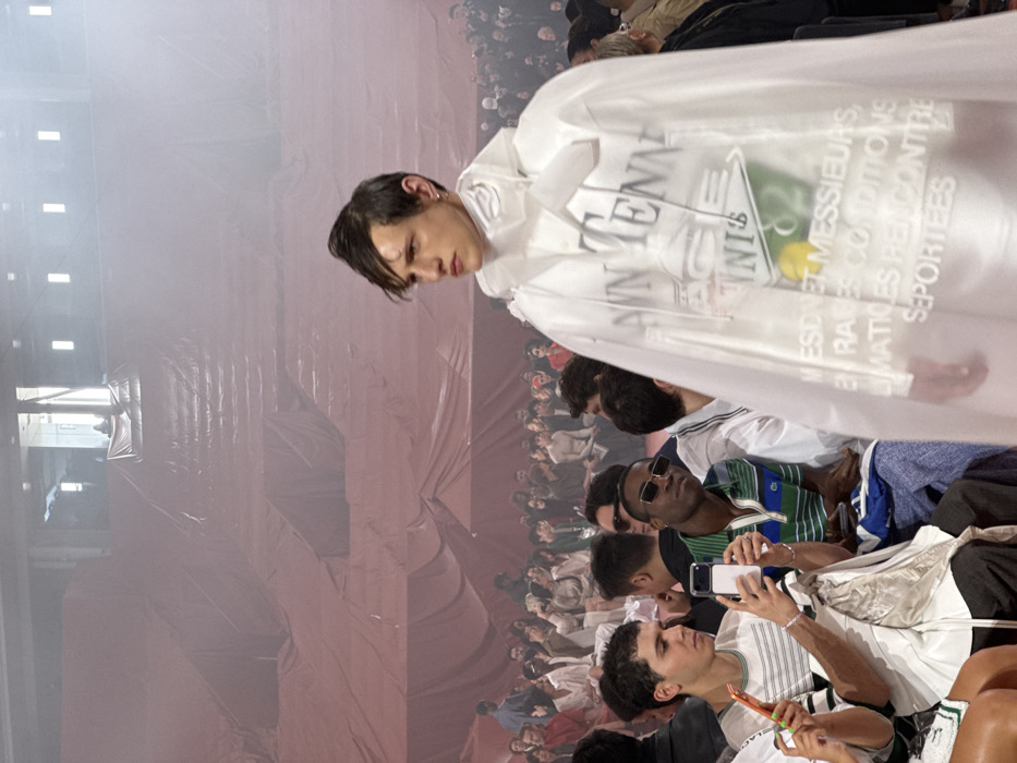 This image captures a fashion event within a large indoor venue, likely the Grand Palais Éphémère in Paris, France. The scene is dominated by a male model walking in the foreground, facing towards the viewer's right. He possesses dark, side-parted hair, an earring in his left ear, and a focused expression. He wears a white high-neck garment, a white hooded top, and an outer clear, hooded PVC-like cape. The cape features black text reading "NN TENNIS," a graphic resembling a green tennis court with a yellow tennis ball, and white French text across the lower portion, partially visible, which reads "MESDAMES MESSIEURS, EN RARES CONDITIONS CLIMATIQUES RENCONTRÉES, SEPORTÉES."
Behind the model and extending into the midground and background, numerous spectators are seated on tiered structures draped in reddish-pink fabric. The atmosphere is hazy or smoky, particularly noticeable in the background, where the audience's forms become less distinct. In the immediate foreground, to the model's left (viewer's right), a young male with short dark curly hair is seated, holding a white smartphone horizontally, seemingly recording the event. He also holds a bright orange object in his left hand. Adjacent to him, another male with dark skin wears dark sunglasses and a green and blue striped polo shirt, observing the model. Further back, more audience members are visible, with some also holding smartphones. The upper background reveals an industrial-style ceiling structure with rectangular light fixtures and a brightly lit opening, possibly an entrance or skylight, at the top of the tiered seating. The overall environment suggests a temporary setup for a large-scale presentation.