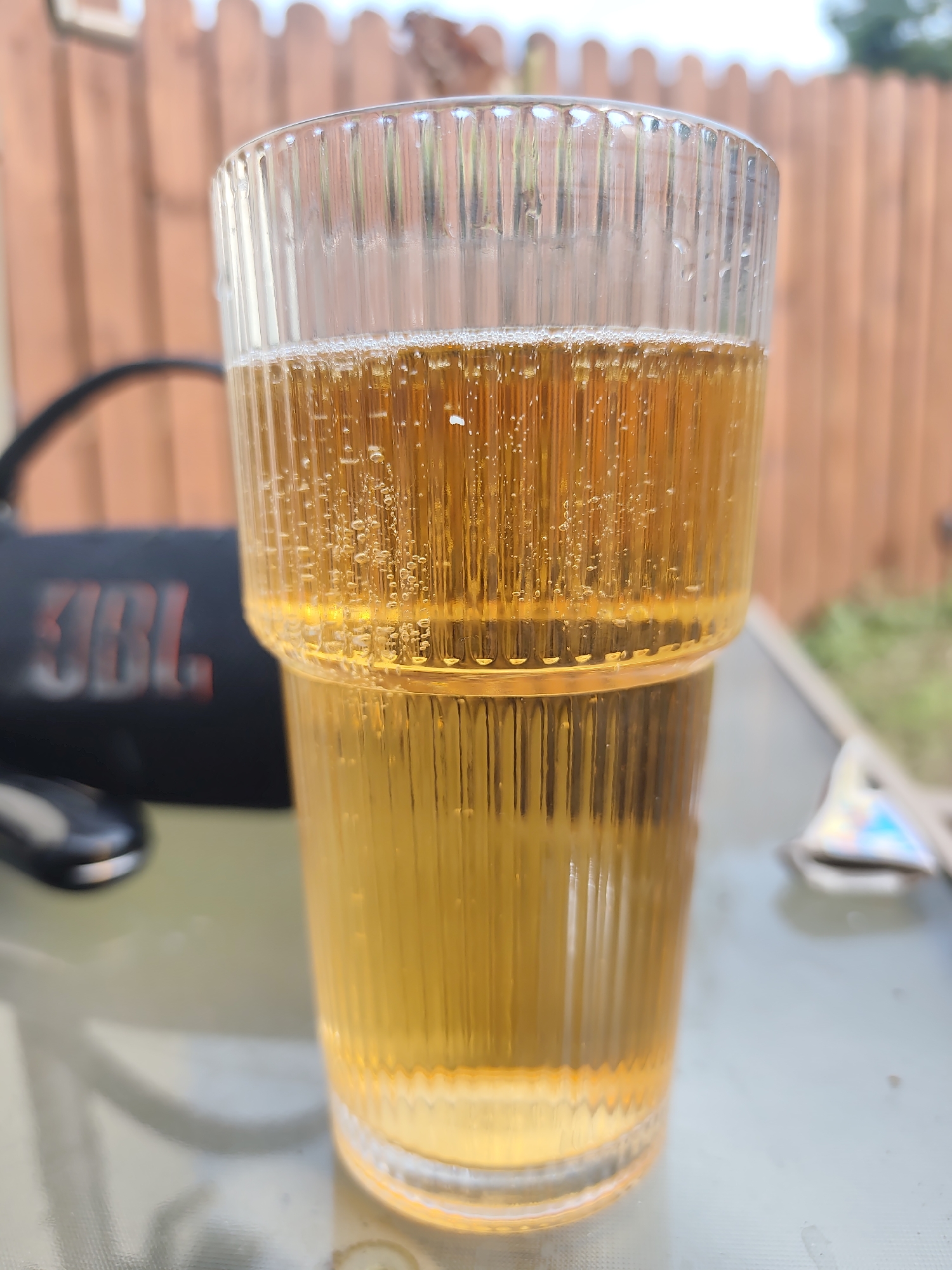 A close-up, eye-level shot shows two stacked clear glasses filled with a golden, carbonated beverage. The glasses have vertical ribbing. The top glass is filled to about two-thirds capacity with a frothy liquid, while the bottom glass is filled to just below its rim. Bubbles are visible within the liquid of both glasses. In the background, slightly out of focus, is a wooden fence and greenery. To the left of the glasses, a black JBL portable speaker is visible, along with a dark, rounded object that appears to be a computer mouse. The scene is set on a reflective surface, likely a table, with another out-of-focus object with a holographic sheen on the right. There are no people present, and the overall setting appears to be outdoors.
This image does not contain any identifiable elements that would allow for its localization in Ballyvary, Ireland.
