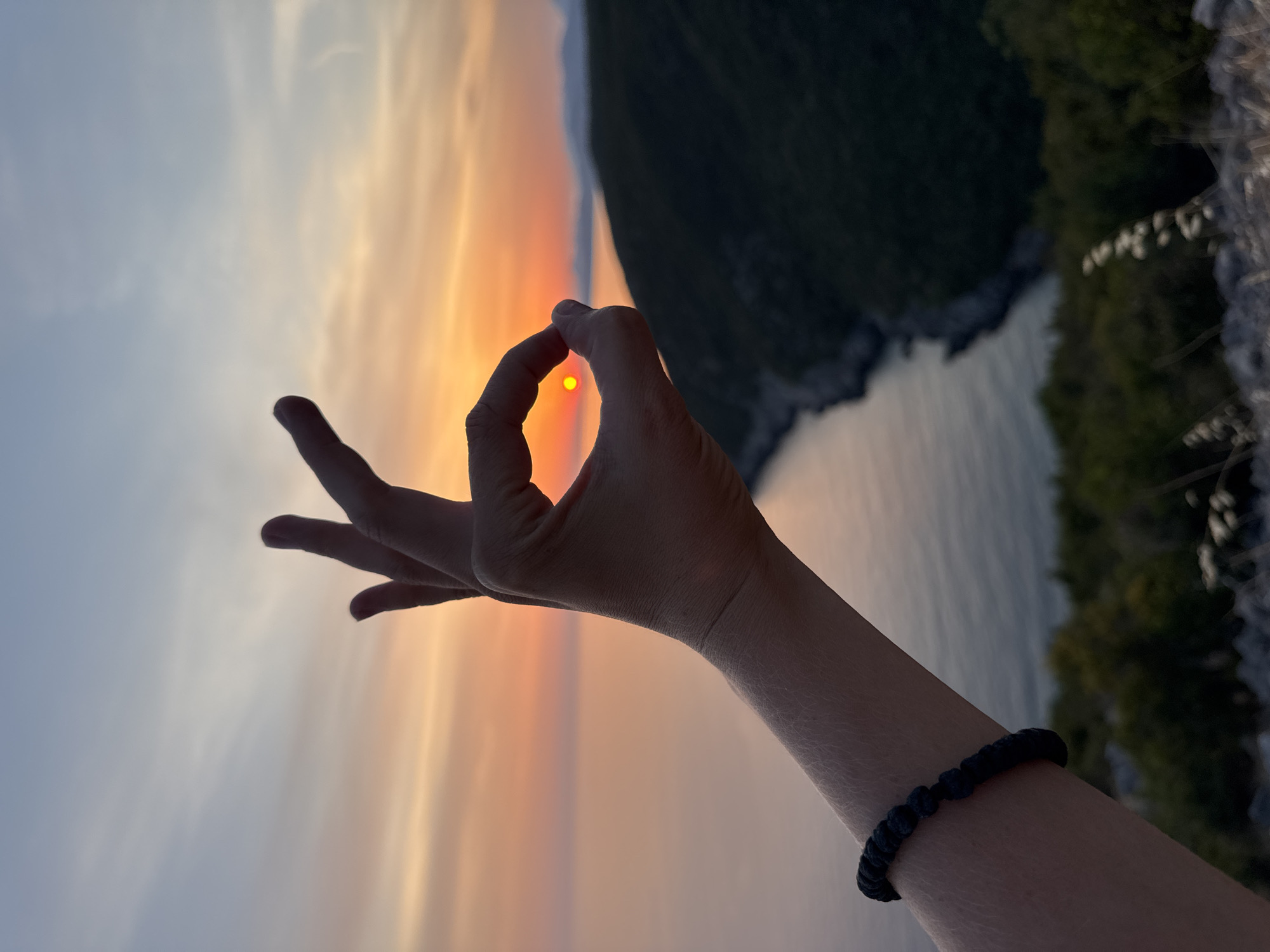 A person's arm and hand form an "OK" symbol in the foreground, silhouetting the sun as it sets over the Adriatic Sea. The arm is adorned with a black beaded bracelet. In the background, a dark, hilly coastline stretches into the distance. The sky transitions from a pale blue at the top to a gradient of orange and pink hues as it meets the horizon. The location is the coast of Zagora, Montenegro.