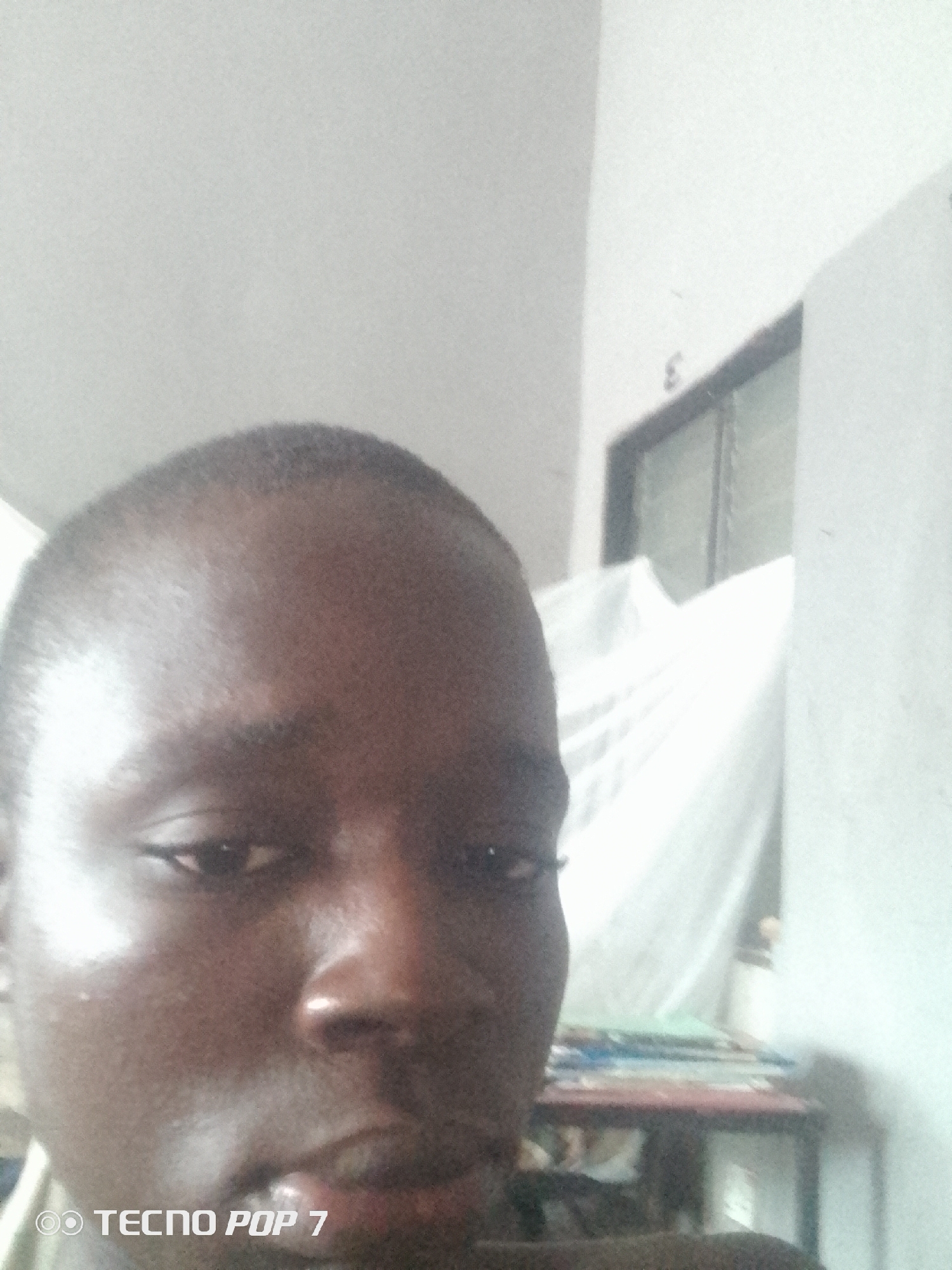 A close-up, low-angle selfie shows the face of a dark-skinned individual, likely a male, with short hair. The individual is looking directly at the camera, with their face filling most of the frame. The lighting suggests an indoor environment, with a white wall and a window visible in the background. A white sheet or curtain is draped behind the individual. A table with some items on it is visible to the right. The bottom left corner of the image contains a watermark indicating "TECNO POP 7". The scene appears to be in a domestic setting within the city of Ipetumodu, Nigeria, suggested by the brand of the phone and the general aesthetic.
