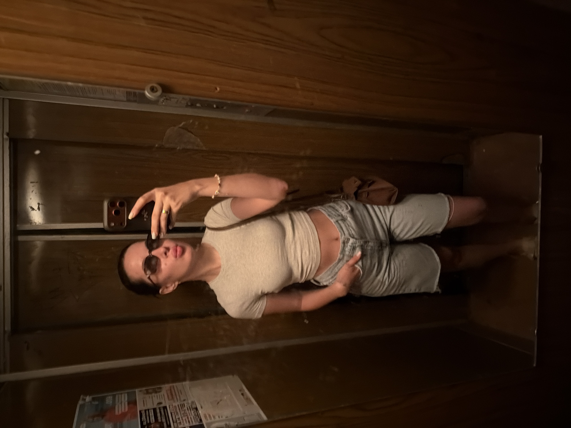 A full-body, mirror selfie shows a woman standing in what appears to be an elevator with wood-paneled walls. She is wearing a light gray, cropped t-shirt, light wash denim shorts with a frayed hem, and a small brown crossbody bag slung over her left shoulder. The woman has on dark sunglasses and is puckering her lips. She is holding her phone in her right hand, angled to capture the reflection. Her left hand is in her pocket. The lighting is dim, casting shadows on the walls.
