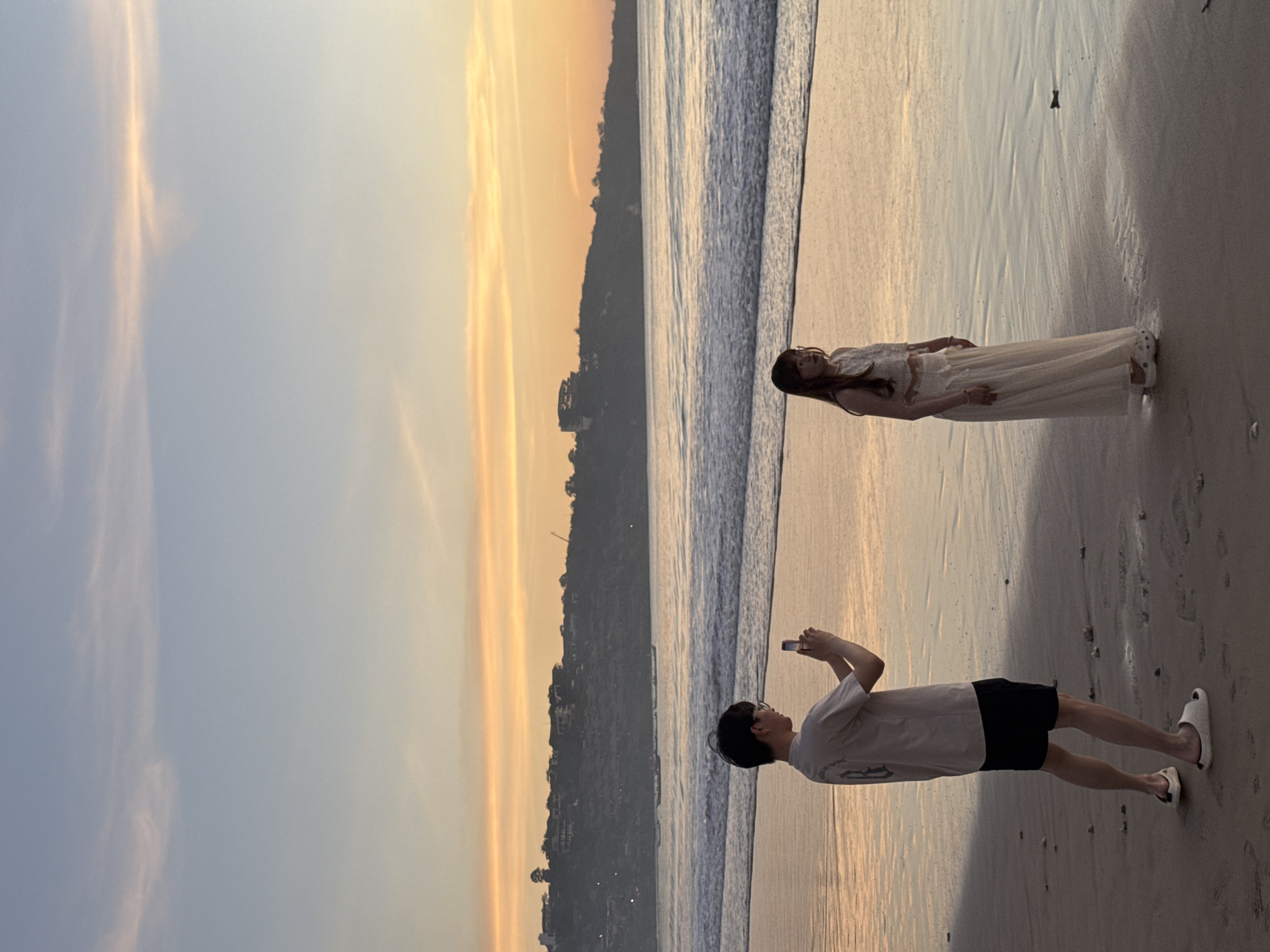 A photograph captures a beach scene at sunset in Kuta Selatan, Indonesia. A woman lies on the wet sand near the ocean's edge, clad in a light-colored, full-length dress. Nearby, a person stands holding a mobile phone, presumably taking a picture of the woman. The individual is wearing a light-colored t-shirt and dark shorts, and is facing the woman. The background features a sunset sky with hues of orange and yellow, contrasted with a darker landmass extending along the shoreline. The beach is wide, with the sand appearing wet and reflective due to the receding tide.