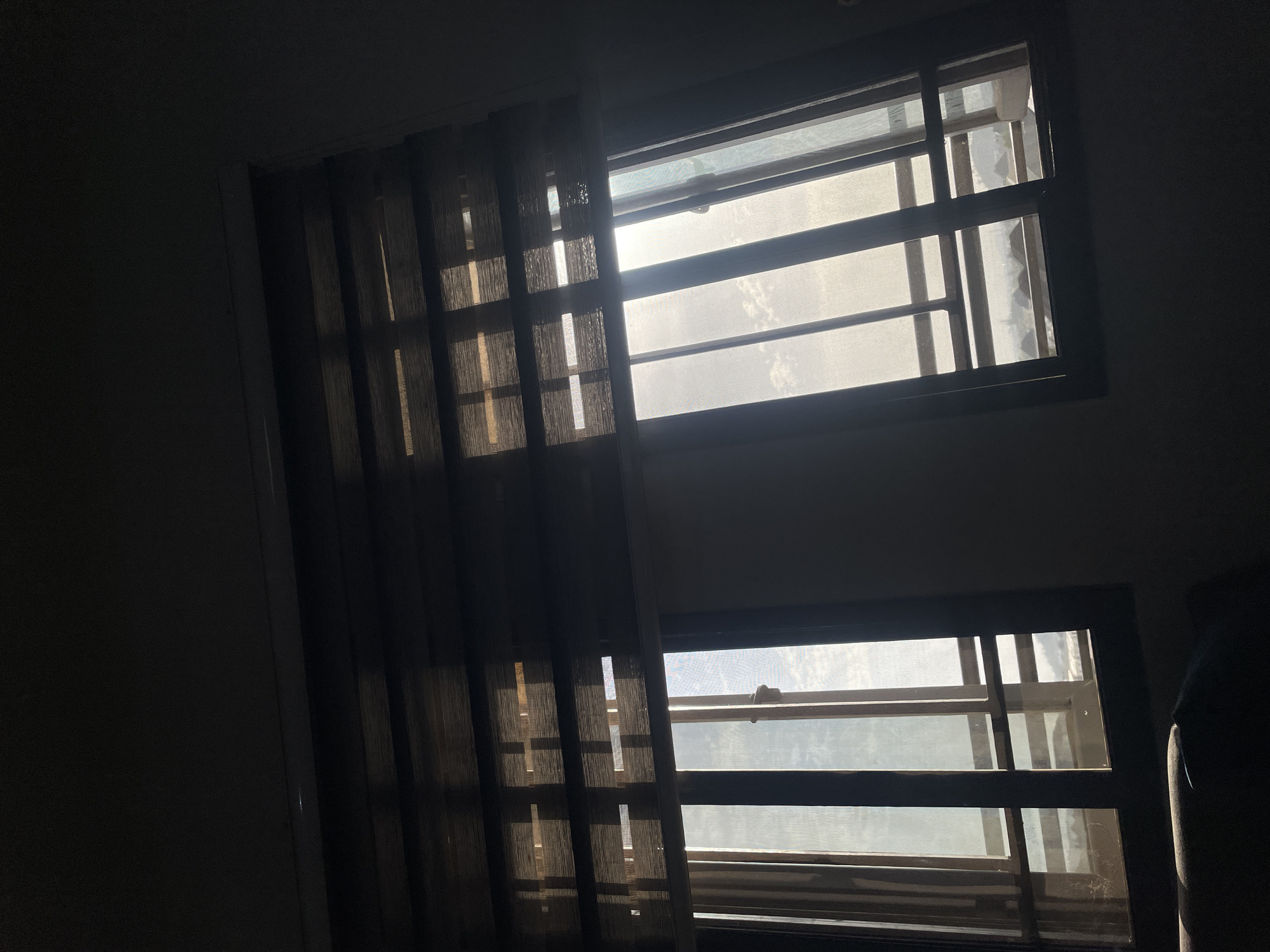 The image presents an interior view looking out through two rectangular windows, set against a dark wall. The room is dimly lit, creating a stark contrast with the bright daylight streaming in from outside.
Each window is framed in a dark material and features horizontal and vertical security grates or bars, visible beyond a semi-transparent mesh screen. This screen diffuses the intense sunlight, making the outside appear as a bright, luminous white space. The top window shows the bright light clearly, while the bottom window, situated below and to the right of the first, reveals a similar bright, diffused light source.
To the left of the windows, dark, semi-transparent vertical blinds or curtains are drawn, partially covering the left side of the top window and almost fully obscuring the wall space to the left of the bottom window. These blinds allow some light to filter through, highlighting their textured fabric and casting subtle light patterns into the dark room.
The scene suggests it is daytime, likely a bright and possibly sunny period, given the intensity of the light from outside. The dark interior could be intentional, perhaps to cool the room or maintain privacy. No people or specific activities are depicted. The architectural style, particularly the security grates on the windows, is consistent with residential buildings in the Abuja, Nigeria region. No visible text is present in the image.