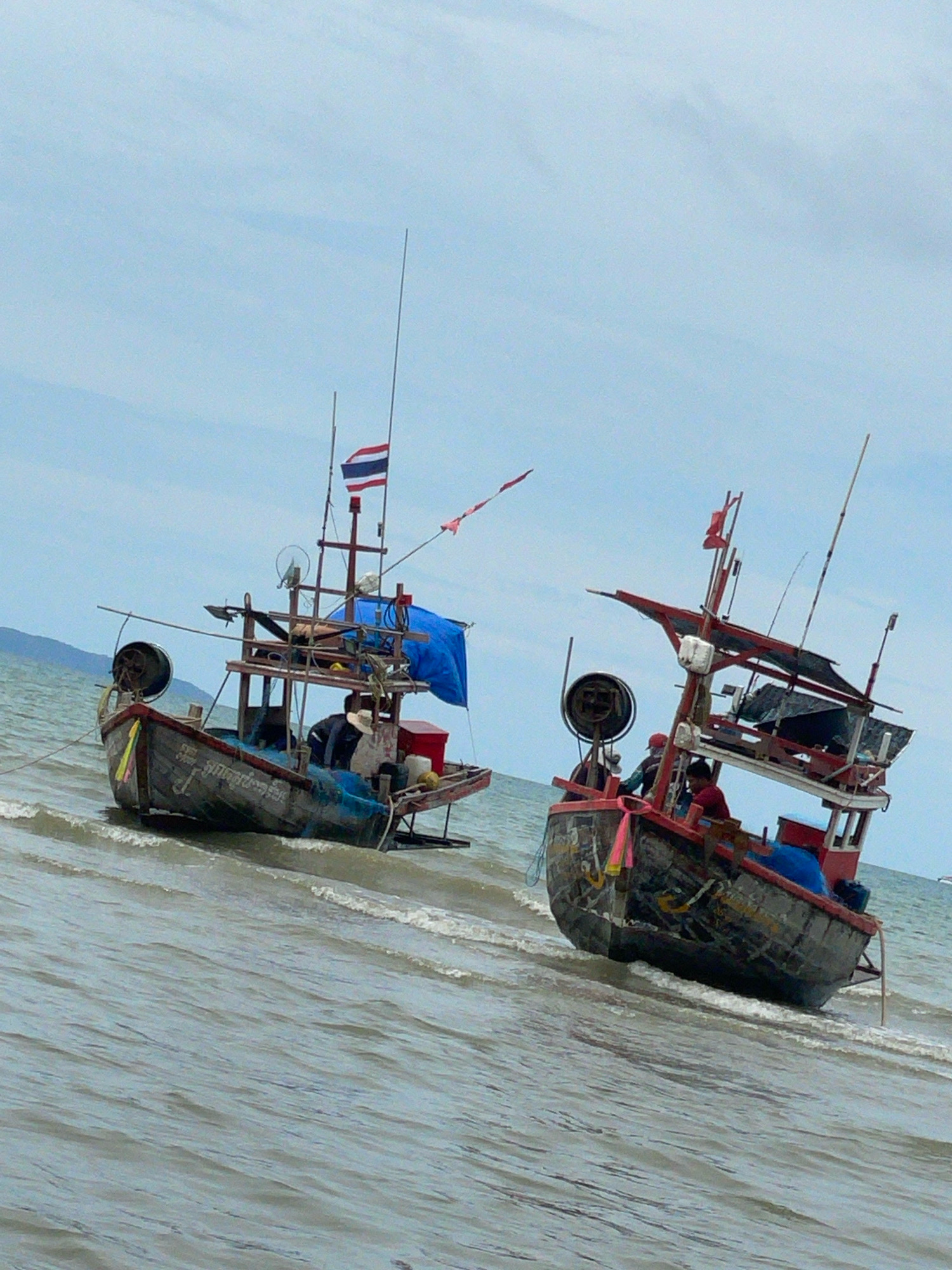 Two small fishing boats are anchored in shallow, choppy water near a beach. The boat on the left is darker, with a weathered hull displaying Thai script. It has a wooden structure with a blue tarp, a Thai flag, and various fishing equipment, including a large spool. A person wearing a hat is visible inside the boat. The boat on the right is red and white, also with a wooden superstructure, a blue tarp, and fishing gear. Two individuals are seated within this boat. The water is a grayish-blue, and the sky is overcast with a pale blue hue. The background shows a hazy coastline with distant hills. The scene appears to be located in or near Ban Na Chom Thian, Thailand, a coastal area known for its fishing activities.