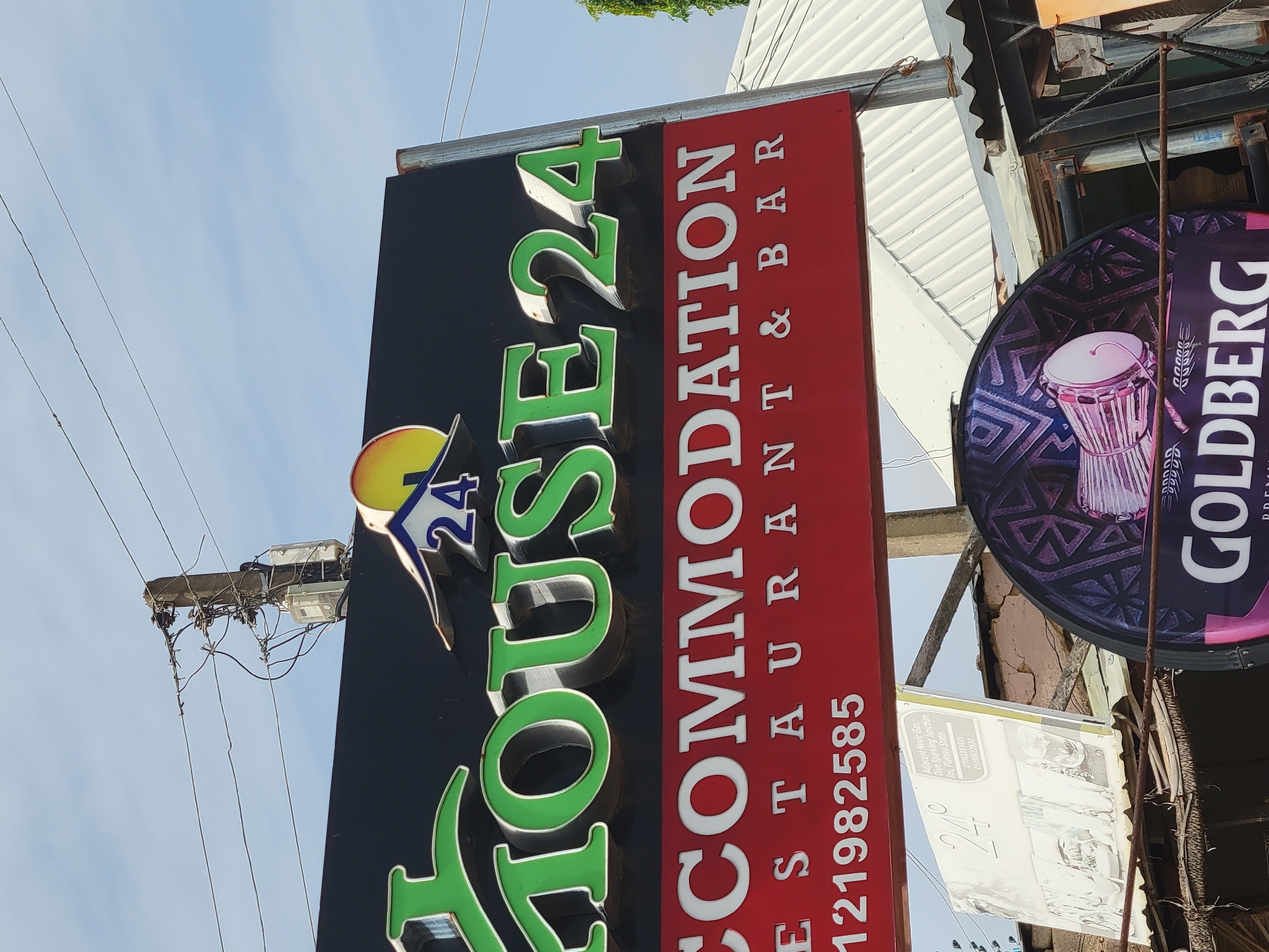 The image captures a street scene in Jos, Plateau State, Nigeria, focusing on signage for businesses. The primary sign is for "HOUSE 24," an establishment offering accommodation, a restaurant, and a bar. Below this, a phone number, "121982585," is displayed. Nearby, there is also a sign advertising "GOLDBERG" beer. A smaller, partially obscured sign mentions "Adjacent Nevic Gas, Diye Sparkling Junction," further pinpointing the location. Contact numbers "07061353435" and "09090622630" are also listed on the smaller sign. The signage is mounted on what appears to be an aged, urban structure. The background includes power lines against a blue sky.