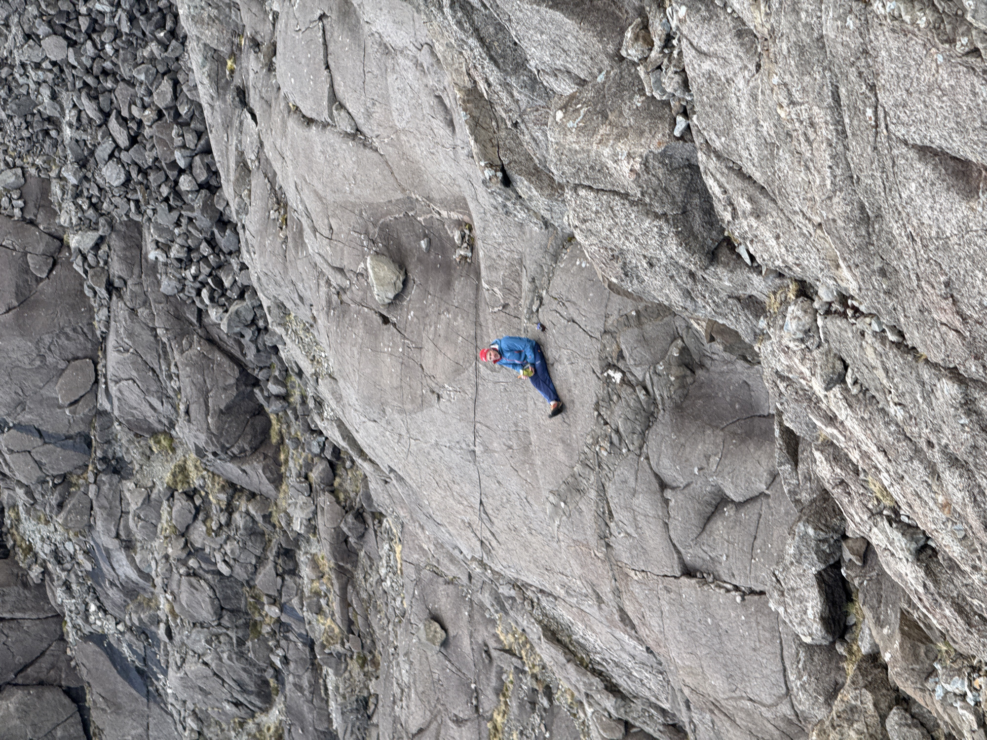 A high-angle view shows a rock climber on a gray rock face. The climber, wearing a blue jacket and red helmet, is situated roughly in the center of the frame. The rock face is jagged and textured. Smaller, loose rocks are visible on the left side of the frame. The location is not specified.