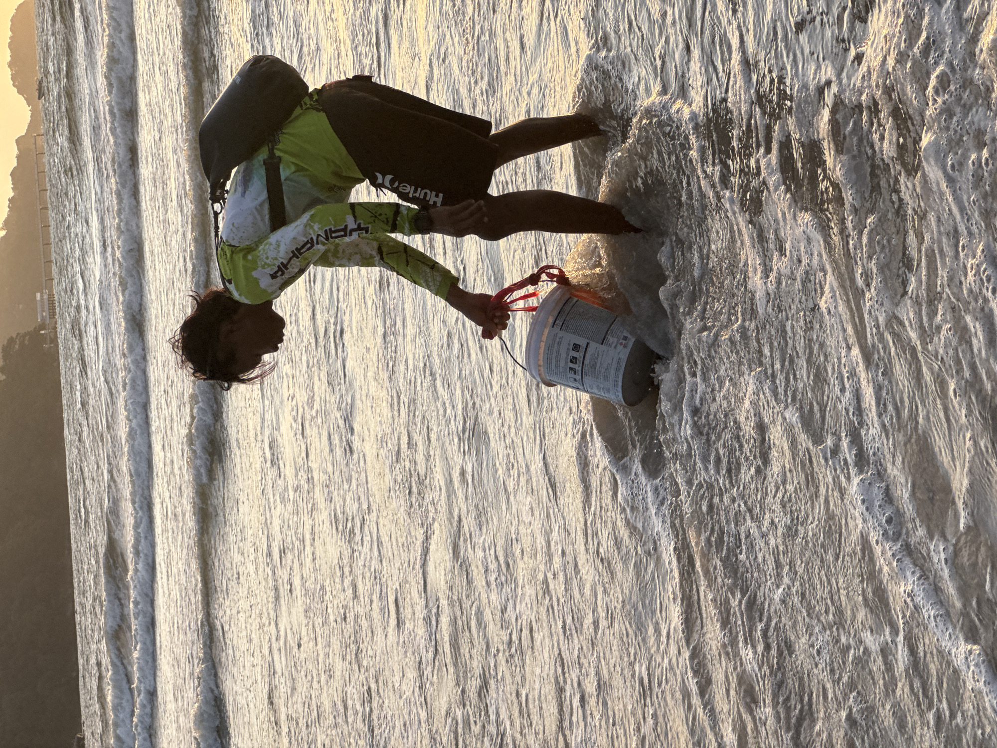In Patong, Thailand, a young person is wading in the shallow surf of a beach during sunset. They are bent over, holding a white bucket with red handles. The bucket appears to be full of something, possibly sand or collected items. The person is wearing a long-sleeved, lime green and white athletic shirt and dark shorts with the "Hurley" logo visible. A dark bag is slung over their right shoulder. The ocean is calm with gentle waves breaking on the shore, reflecting the warm glow of the setting sun. In the background, a distant pier and a boat can be seen, with hills covered in vegetation silhouetted against the bright sky.