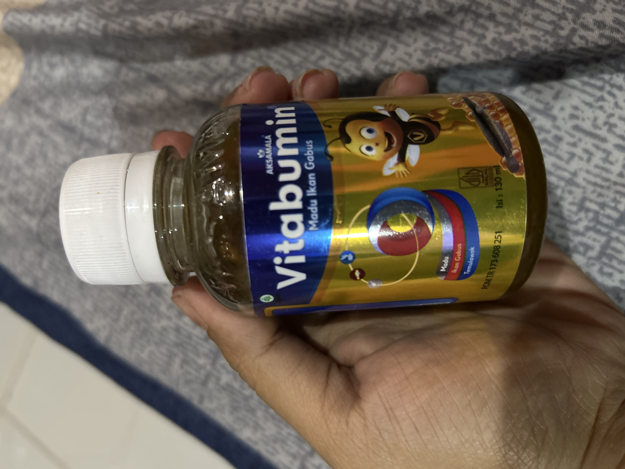 A hand is holding a bottle of Vitabumin Madu Ikan Gabus, a supplement product. The bottle has a white cap and a label with blue and gold coloring. The label features text including "Vitabumin", "Madu Ikan Gabus", "Temulawak", and "POM TR 173 608 251". There is also a cartoon image of a bee on the label. The background appears to be a patterned fabric, possibly a blanket or bedspread. The lighting suggests an indoor setting. The context of Depok, Indonesia, is not directly discernible from the visual information in the image itself.