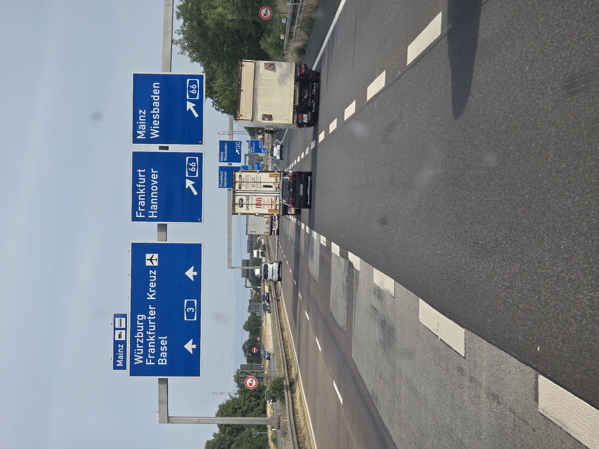 An overhead view captures a highway scene with multiple lanes of traffic under a hazy sky. Prominent blue highway signs provide directional information. One large sign indicates "Würzburg," "Frankfurter Kreuz," and "Basel," with arrows pointing straight ahead and a number "3" enclosed in a white rectangle. To the right, two other signs display "Frankfurt Hannover" and "Mainz Wiesbaden," each with an arrow indicating a turn and the number "66" in a white rectangle. Several vehicles, including cars and trucks, are visible on the highway. The road surface shows signs of repair with patched areas. The environment is urban with trees and vegetation visible along the roadside and in the distance, suggesting the location is near Breckenheim, Germany, given the highway signs.