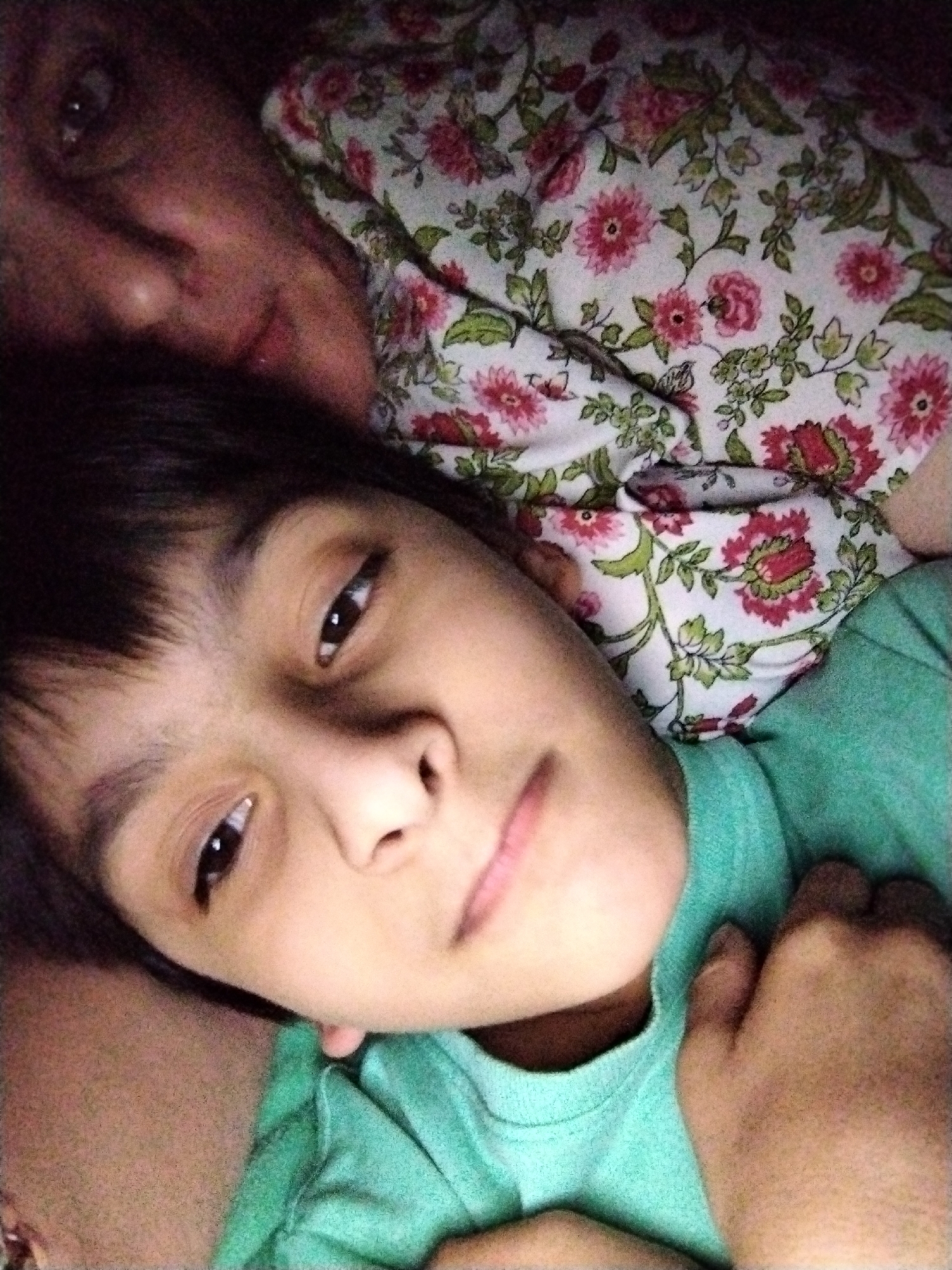 The image presents a close-up, portrait-oriented view of two individuals, an adult and a child, positioned horizontally across the frame. Both subjects appear to be lying down, looking towards the camera.
The child is in the foreground, occupying the lower half of the image. They have dark hair and dark eyes, with a neutral expression and slightly parted lips. They are wearing a plain, light teal short-sleeved t-shirt. Their left hand, with fingers slightly curled, rests near their chest.
Behind and above the child is an adult, partially visible. The adult's right eye, the bridge of their nose, and a portion of their mouth are discernible. Their expression is neutral, and they also appear to be looking towards the camera.
A fabric with a floral pattern is visible between the two individuals and extending behind the adult's head. The pattern consists of red and pink flowers with green leaves on a light background. The lighting in the scene is dim and warm, contributing to a soft, slightly grainy quality. The background behind the adult is dark and indistinct.