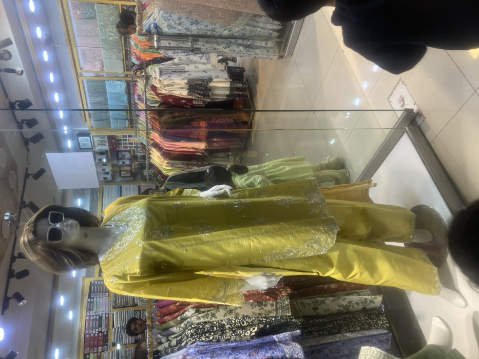 The image depicts the interior of a clothing retail store located in Quetta City Tehsil, Pakistan. In the foreground, a female mannequin stands centrally, dressed in a bright yellow, embroidered long-sleeved kurta and matching wide-legged trousers. The mannequin has brown hair and wears large white-framed sunglasses.
Behind the mannequin and visible through a clear glass partition, the store environment is well-lit with overhead track and recessed lighting. Numerous clothing racks display a variety of women's traditional Pakistani garments in various colors and patterns. Upper shelves in the background contain folded textile merchandise.
Several individuals are partially visible: an adult male's head is seen in the upper-left background, positioned behind a clothing rack. A small child, wearing a light green dress, is visible as a reflection in the glass partition to the right of the central mannequin, facing away from the viewer. The tops of two other children's heads are visible in the bottom-left and bottom-right foreground, indicating their close proximity to the camera. The floor consists of light-colored tiles.