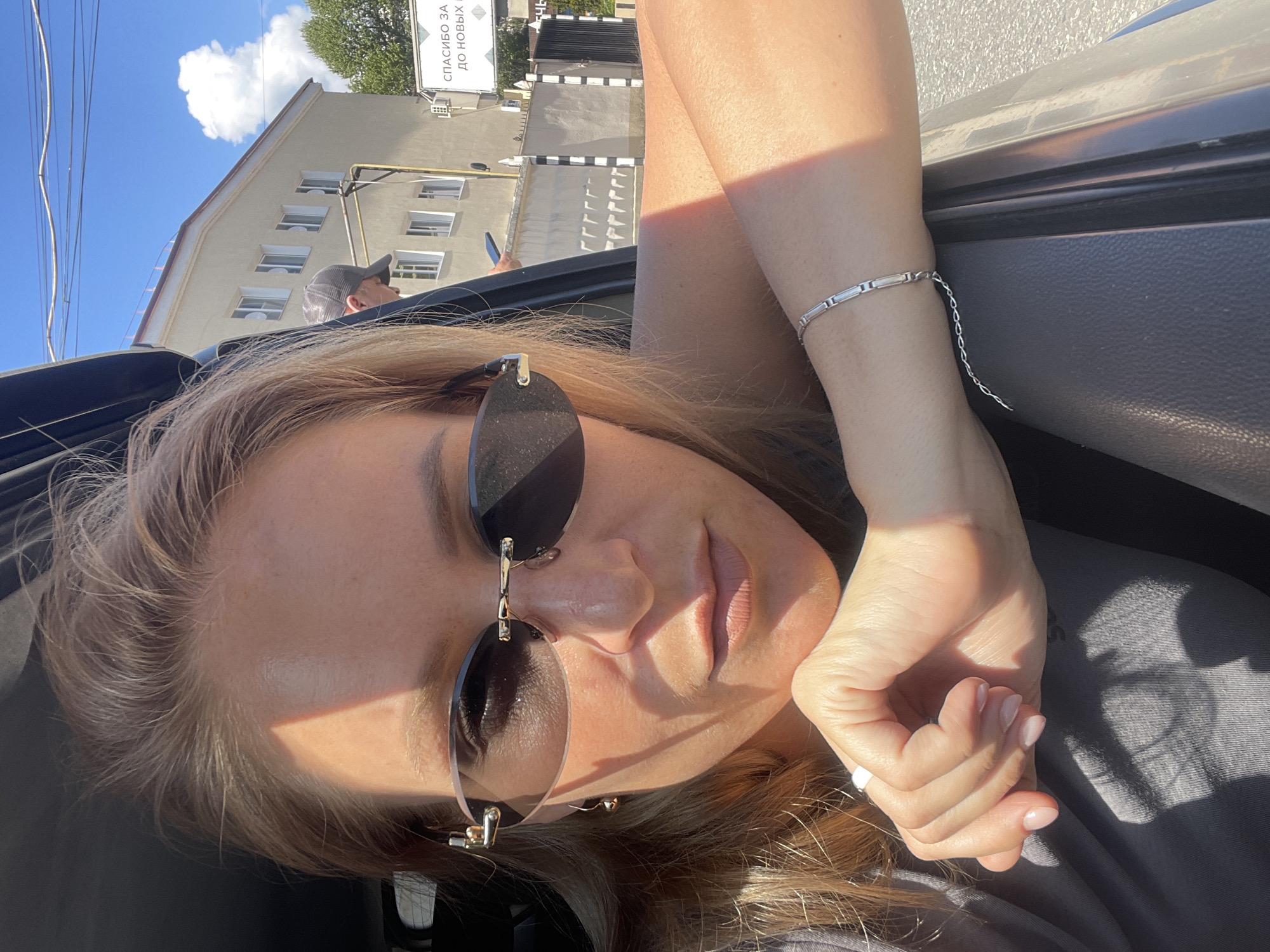 A woman in sunglasses is in a car in Sochi, Russia, on a sunny day. Her arms are crossed in front of her. She is wearing a bracelet on her left wrist. In the background, a tall, tan building is visible, with a sign in Cyrillic characters that reads "Спасибо за до новых," visible above it. Another person is seen behind the car.
