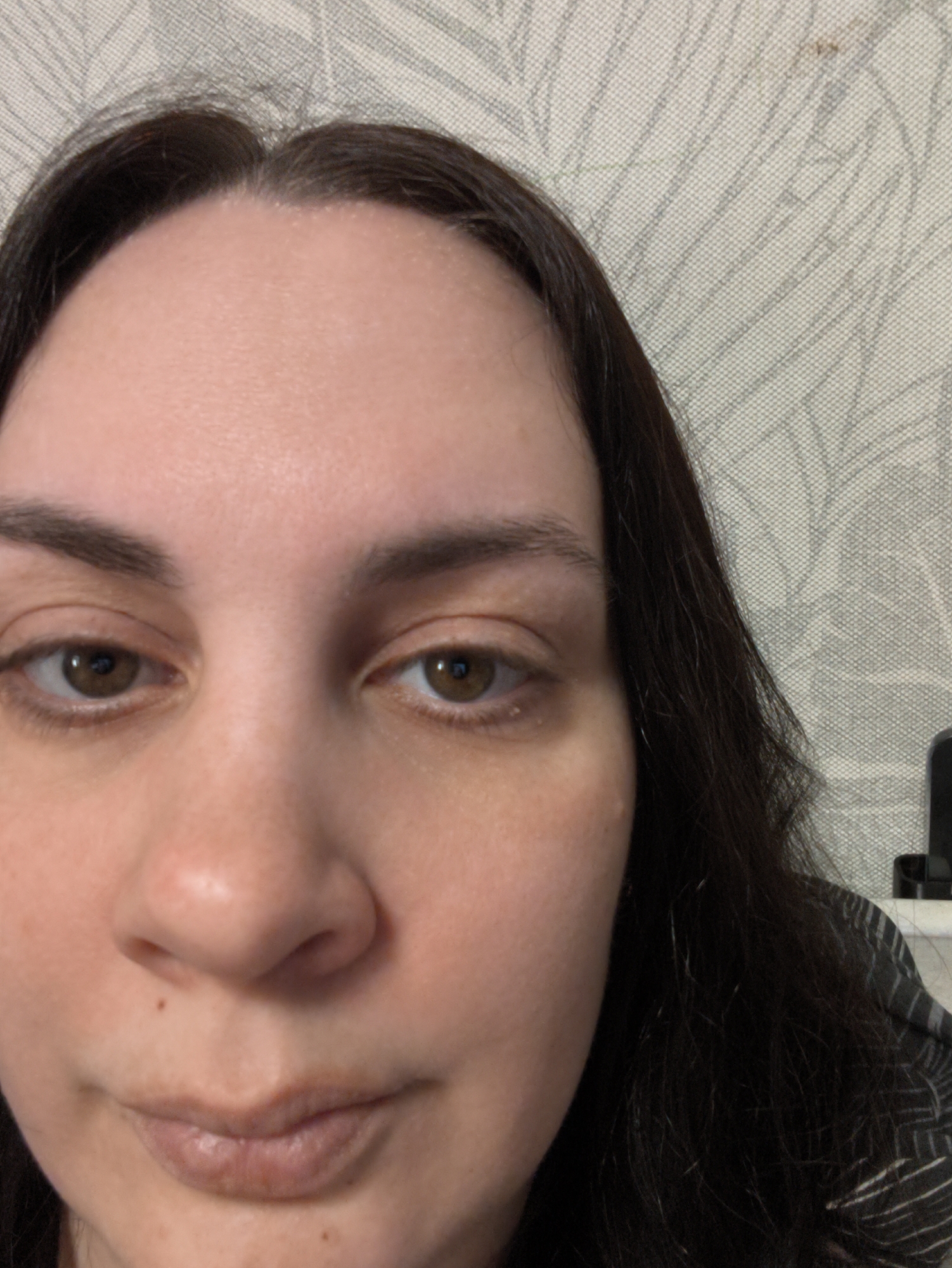 A close-up, indoor photograph shows the face of a woman with dark brown hair. The background is a textured wall with a light gray pattern reminiscent of leaves. The woman has fair skin, brown eyes, and slightly parted lips. A small mole is visible on her left cheek and above her upper lip. The left side of her hair partially obscures the right side of the frame. Part of a black object is visible in the lower right corner. No evidence suggests the image was taken in Chaikovsky, Russia.