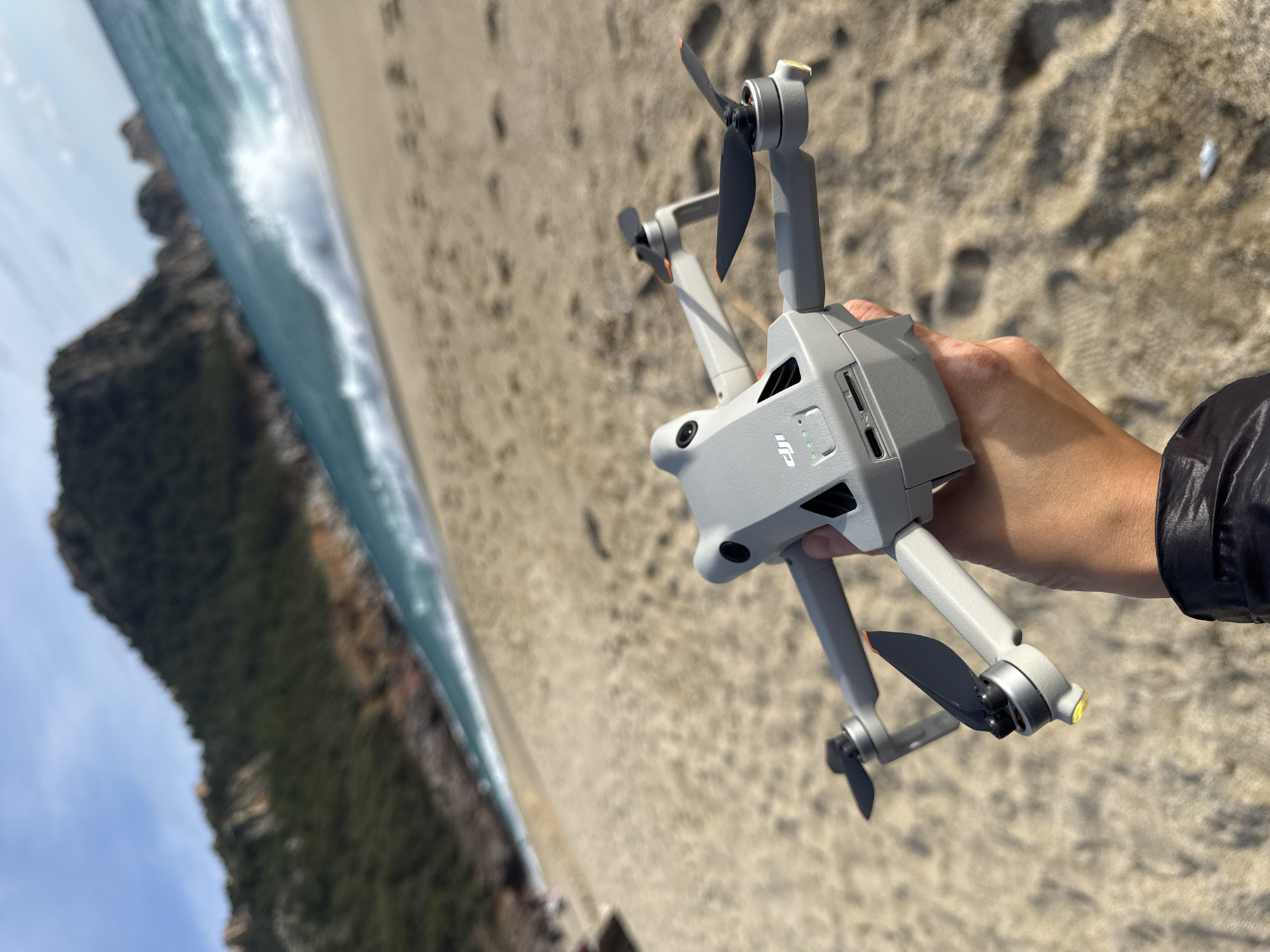 A grey DJI Mini 3 Pro drone is prominently held in the air by a human hand, with four green indicator lights illuminated on its top surface, suggesting it is powered on. Two propellers with black blades and orange tips are visible. The hand, displaying a light to medium skin tone, is partially obscured by the sleeve of a dark, possibly synthetic, jacket. The drone's underside reveals a USB-C port and an SD card slot.
The background depicts a sandy beach with visible footprints. Beyond the sand, a body of turquoise-blue water features white breaking waves. In the far distance, steep, verdant mountains covered in trees and scattered rocky outcrops rise against a partially cloudy blue sky. This coastal environment is consistent with Cleopatra Beach or a similar area along the Mediterranean coast in Alanya, Turkey, characterized by its sandy shores, clear waters, and mountainous hinterland.