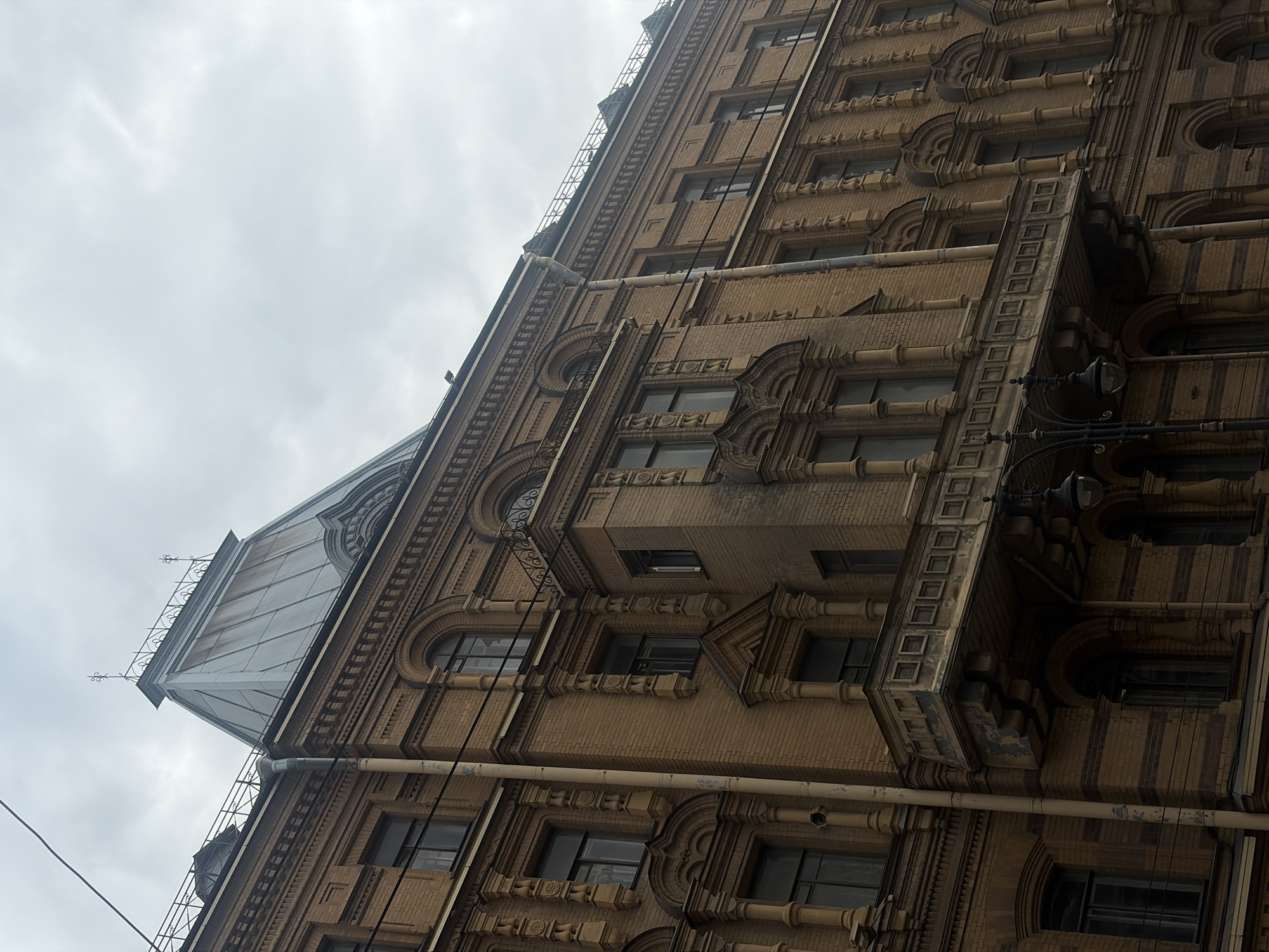 The image shows a low-angle view of a tall, historic building in Saint Petersburg, Russia, under an overcast sky. The building's facade is constructed of brick and adorned with intricate architectural details, including rows of small, square indentations, decorative moldings, and arched window frames. Several windows are visible, appearing dark and reflecting the cloudy sky. A distinctive pitched roof section covered in metal panels is also present, topped with decorative ironwork. Various pipes and conduits run along the exterior of the building. The overall impression is of an aged, grand structure on a day with diffused light, typical of Saint Petersburg's often cloudy weather. No people or specific activities are depicted, and no text is visible.