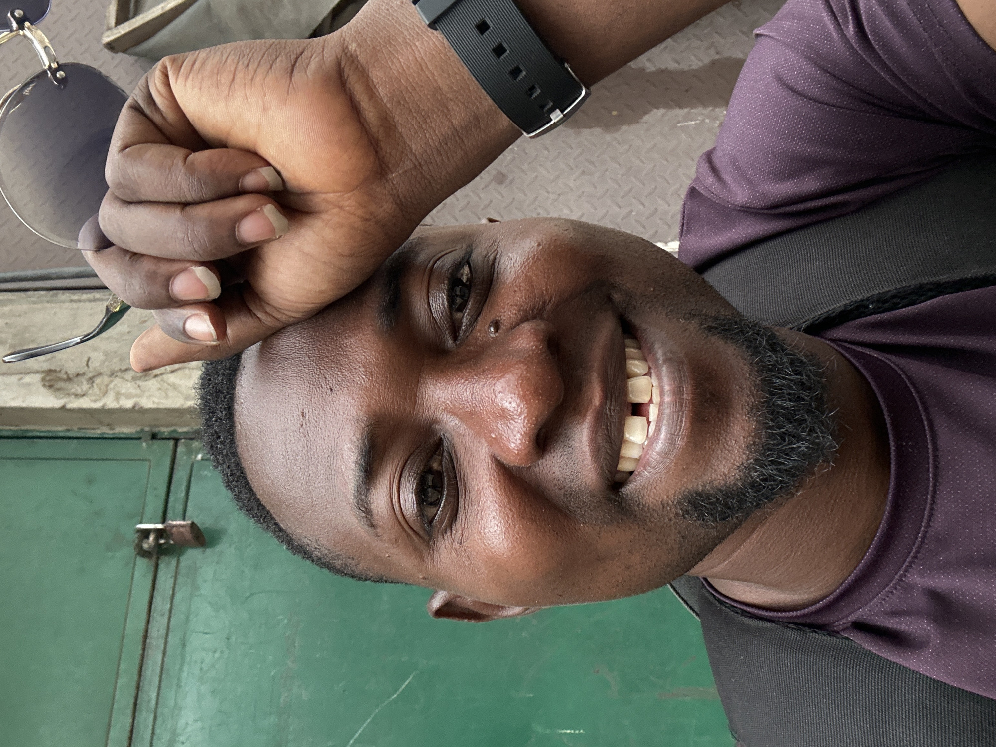 An overhead, rotated close-up selfie captures a smiling man, likely of African descent, with dark skin, short dark hair, and a neatly trimmed goatee beard. He is looking directly at the camera with dark brown eyes and a wide, joyful smile that reveals his teeth. A small piercing adorns his right nostril.
His left hand, holding a pair of rimless sunglasses with dark lenses, rests on his forehead. His fingers and fingernails are visible. On his right wrist, a black smartwatch with a textured strap is prominent. He is dressed in a dark purple short-sleeved shirt with a subtle, dotted texture, possibly athletic wear, and a black undergarment or strap is visible beneath it.
The background elements suggest an indoor or covered outdoor setting. To the upper right, a light grey metallic surface with a diamond plate pattern can be seen. To the lower left, a dark green metallic door or cabinet is visible, complete with a latch mechanism. The overall impression is one of candid happiness. No text is visible in the image.