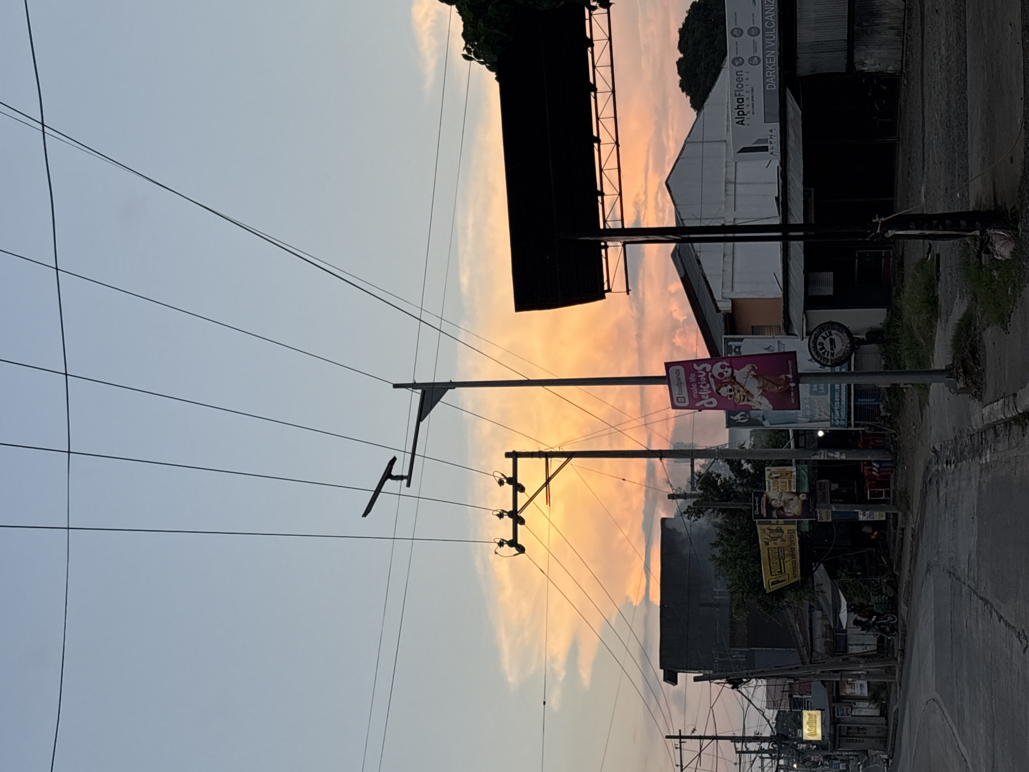 The image captures a street scene in Davao City, Philippines, during a sunset. The sky is painted with vibrant orange and pink hues due to the setting sun, illuminating the clouds. Prominent in the foreground are several utility poles with numerous overhead wires, suggesting a developed urban area. A street with a slight curve is visible, lined with various establishments, including a building with a dark, cantilevered roof, possibly a commercial space or a carport. Signs for "AlphaFloen Financial" and "DARKEn VULCANIZIN" are visible on one of the buildings. In the middle ground, there's a cluster of buildings, some appearing older and more weathered, with a small canopy structure. A few motorcycles are parked near one of the establishments, and two figures are standing nearby. A large billboard featuring a cartoon character and the "foodpanda" logo is also present. The overall impression is of a typical urban environment in the Philippines as the day transitions into evening.