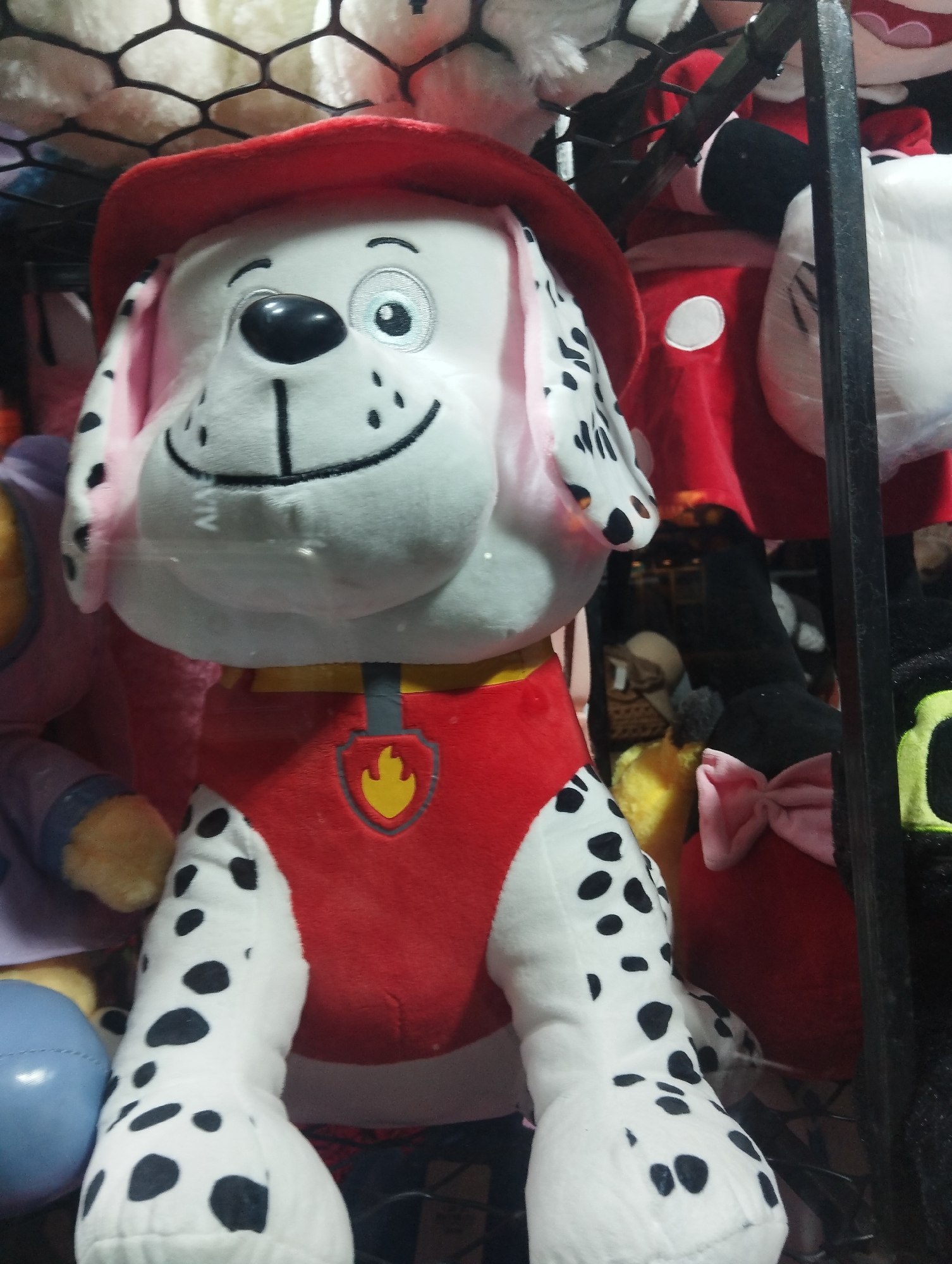 A plush toy of Marshall from Paw Patrol is partially visible in a store display. The toy is a white Dalmatian with black spots, wearing a red fireman's hat and vest. The vest has a yellow fire emblem on it. The toy has a stitched smile and a single visible embroidered eye. Other plush toys are visible in the background. The location is most likely in a commercial establishment within El Valle del Espíritu Santo, Venezuela.