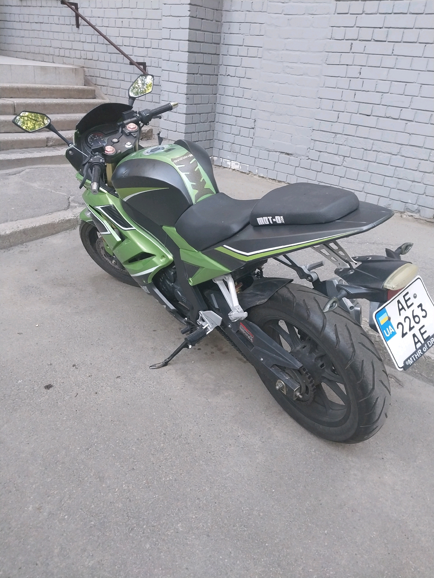 The image shows a green and black sport motorcycle parked on an asphalt surface. The motorcycle is positioned at an angle, facing away from the viewer and towards the right. A license plate with the characters "AE 2263 AE" and a Ukrainian flag emblem is visible on the rear of the motorcycle. The motorcycle has a black seat and is leaning on its kickstand. In the background, there is a brick wall and a set of concrete stairs leading up to a building. The environment suggests an urban setting. Based on the license plate information and visual cues, this scene is likely located in the city of Dnipro, Ukraine. There are no people visible in the image.