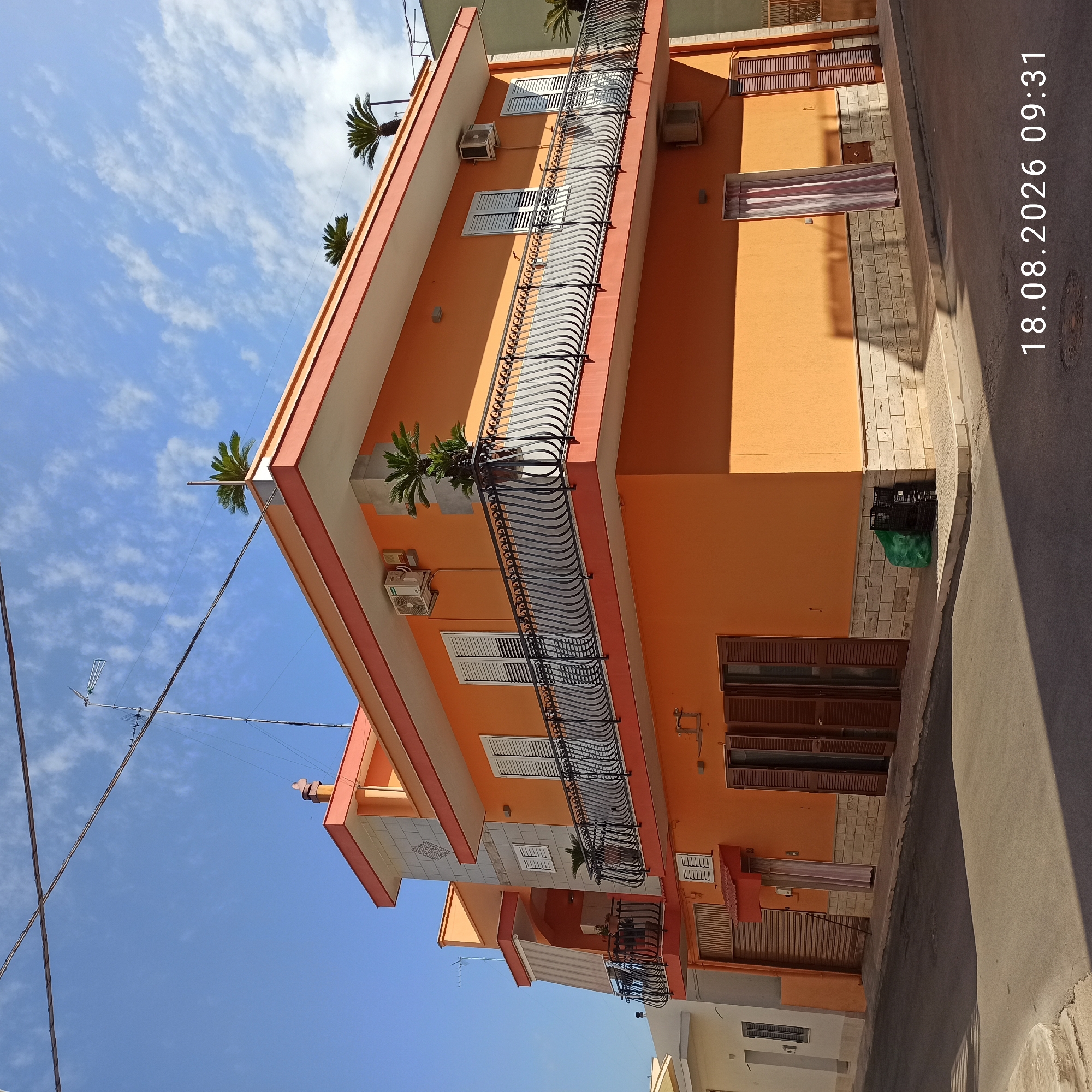 The image depicts a corner of a multi-story residential building painted orange with red trim, located in Bitonto, Italy. The building features balconies with decorative black railings and shutters on the windows. An air conditioning unit is mounted on the exterior wall. In front of the building, on the sidewalk, are a green trash bag and two black plastic crates. The street in front of the building is paved and partially shaded by the building itself. The sky above is blue with scattered white clouds. Power lines and an antenna are visible against the sky. The timestamp "18.08.2026 09:31" is overlaid on the image.