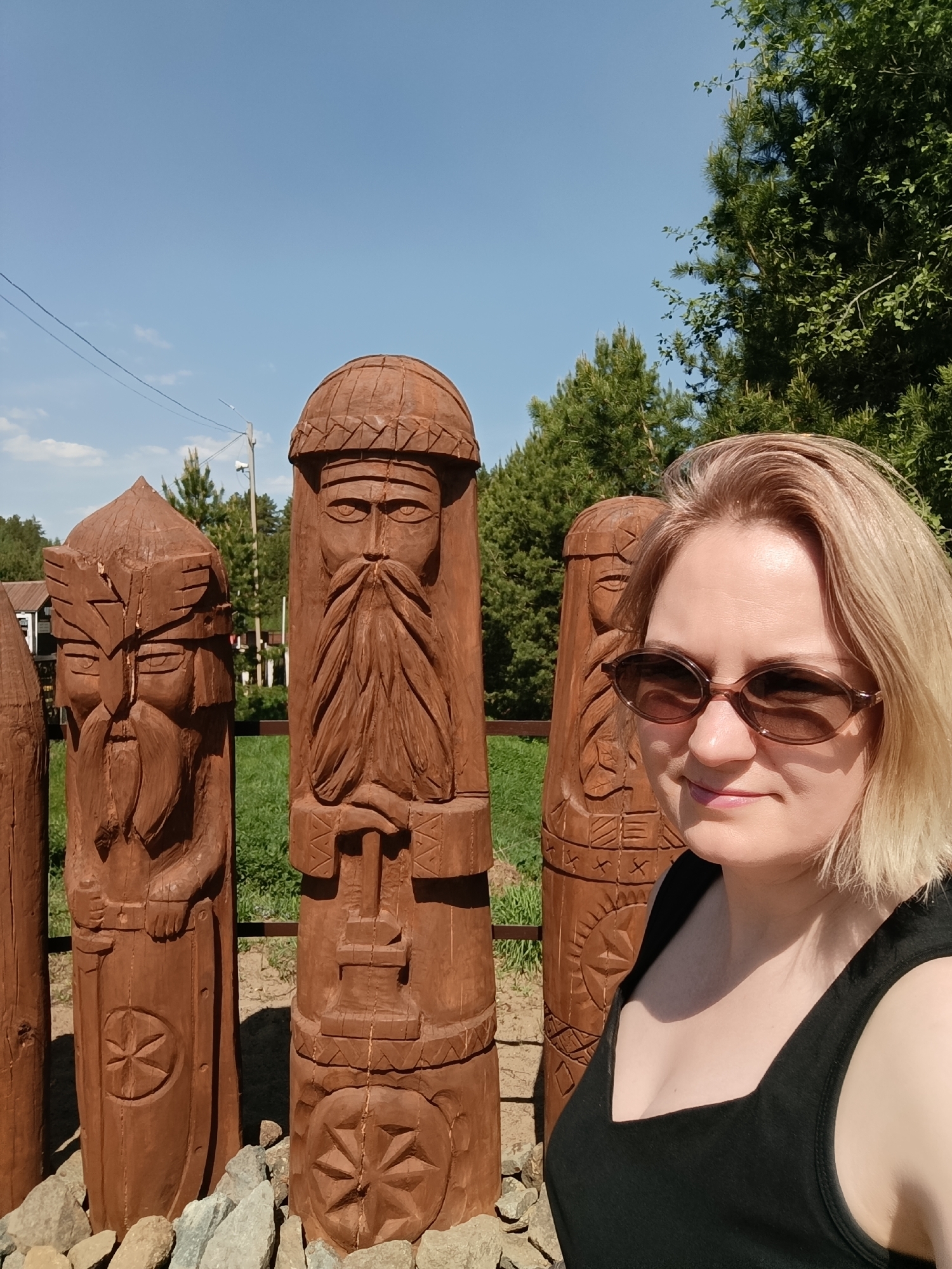 The image depicts a woman in a black tank top and sunglasses standing next to several wooden statues. The statues are carved with faces and appear to be depictions of deities or mythical figures, exhibiting intricate detail. The ground around the statues is covered with small rocks. In the background, there are trees, grass, and a house. The scene is set outdoors under a clear blue sky. Based on the location provided, the photo was likely taken in the city of Prikamskii, Russia.