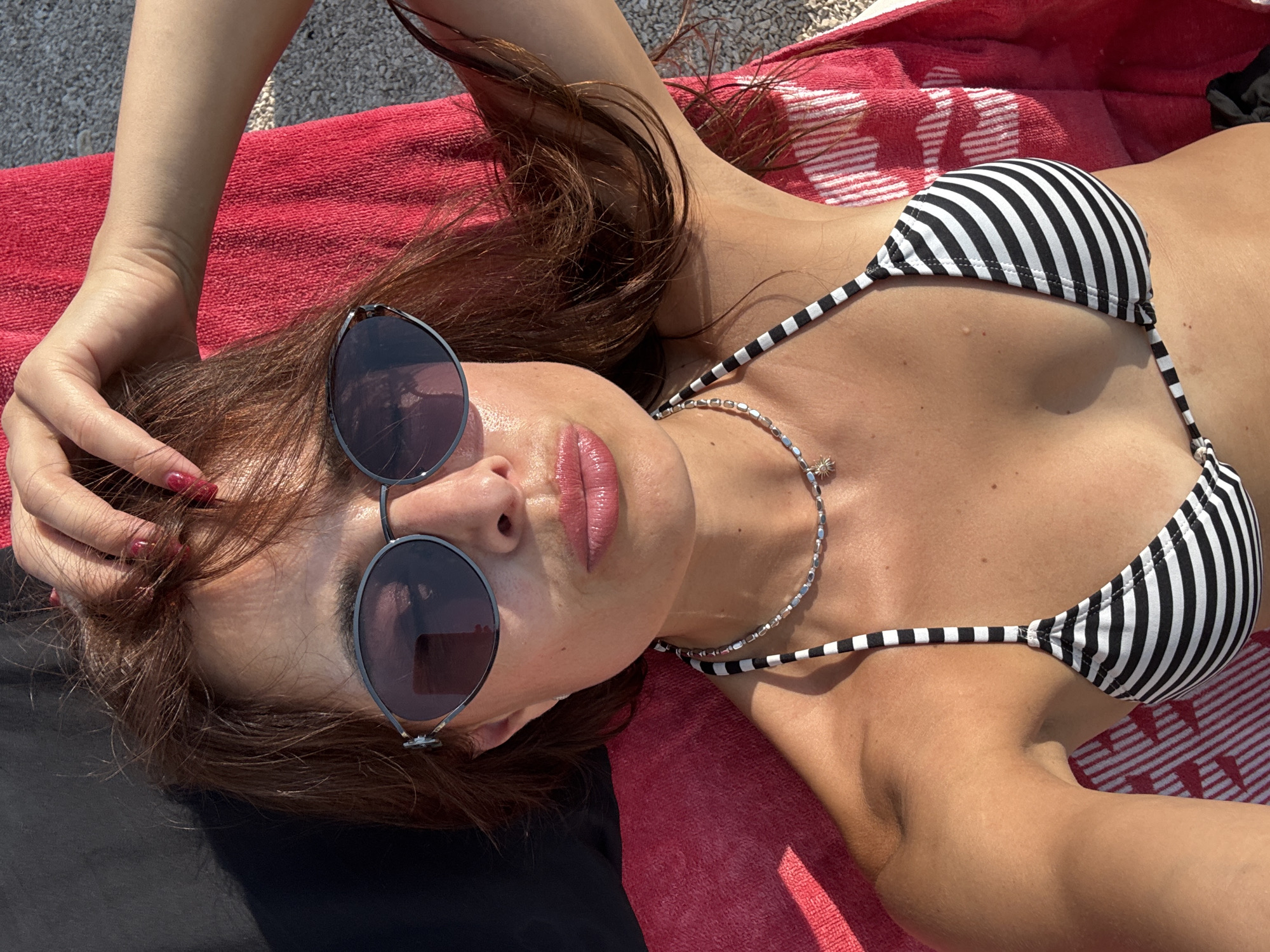 The media file shows a close-up, overhead shot of a woman lying on a red and white striped towel. She is wearing a black and white striped bikini and sunglasses. Her hand is in her hair, and her lips are parted slightly, suggesting a relaxed or sensual pose. The lighting suggests it is daytime, likely sunny, as indicated by the strong shadows and the glint on the sunglasses. The texture of the towel and the woman's skin are clearly visible. There is no visible text in the image. The overall impression is that of a leisurely moment spent outdoors, possibly at a beach or poolside.
