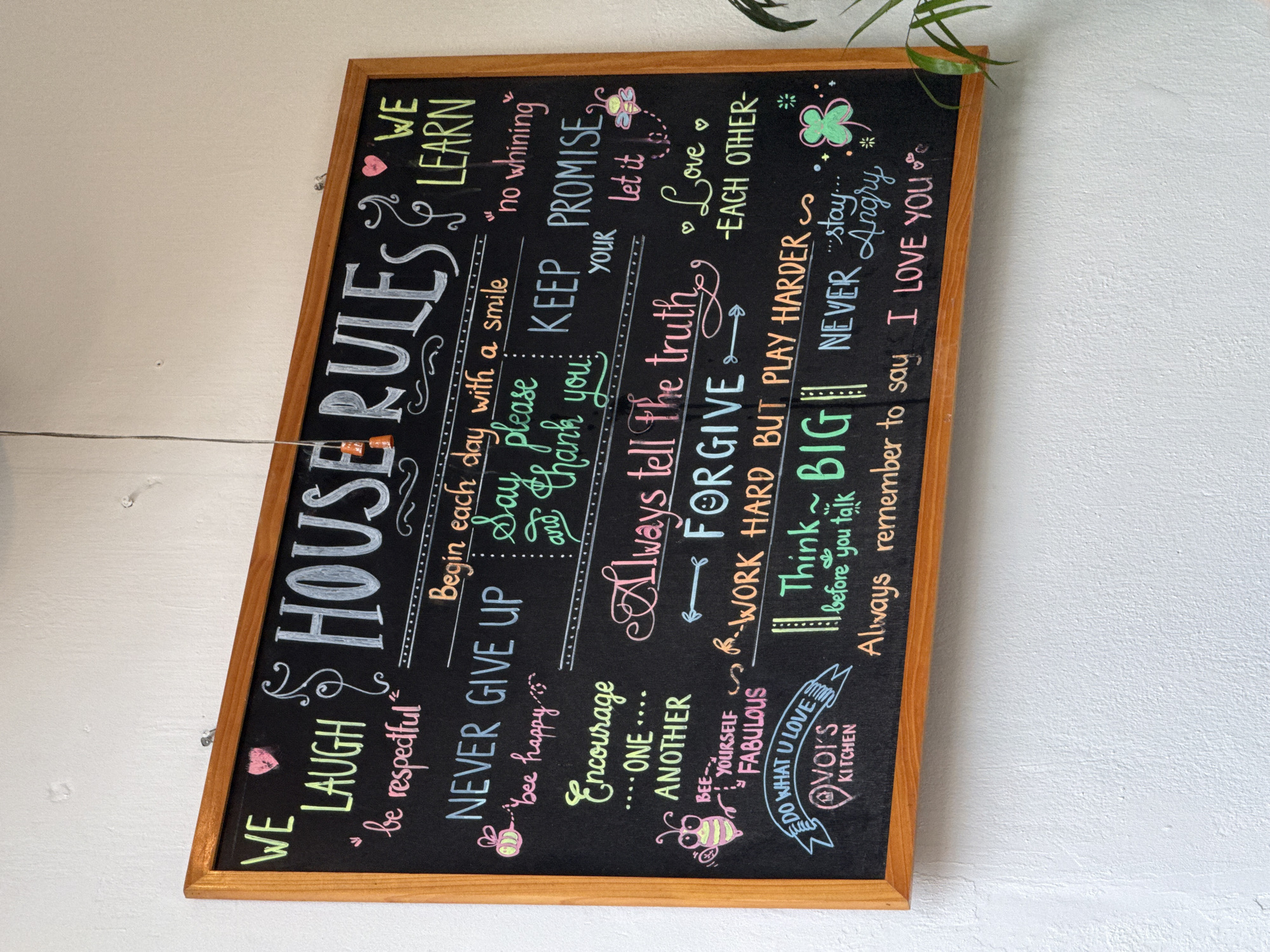 Here is a detailed description of the image:
**Scene:**
The image shows a chalkboard hanging on a white wall. A few plant leaves are visible in the top right corner. The chalkboard is framed in brown wood.
**Text on the chalkboard (house rules):**
* WE LAUGH "be respectful"
* HOUSE RULES
* WE LEARN
* Begin each day with a smile
* NEVER GIVE UP "bee happy"
* Say Please and Thank You
* KEEP PROMISE "no whining"
* YOUR Let it
* Encourage ONE ANOTHER
* Always tell the truth
* Love Each Other
* BEE YOURSELF FABULOUS
* FORGIVE Work Hard But Play Harder
* Think Big before you talk
* Never stay angry
* Always Remember to say I Love You
* Do what you Love. VOI'S Kitchen
**Objects:**
* Chalkboard
* Brown frame
* Plant Leaves
* Wall
**Colors Used:**
* Black (Chalkboard background)
* White, Yellow, Pink, Green, Light Blue (Text)
* Brown (Frame)
* White (Wall)