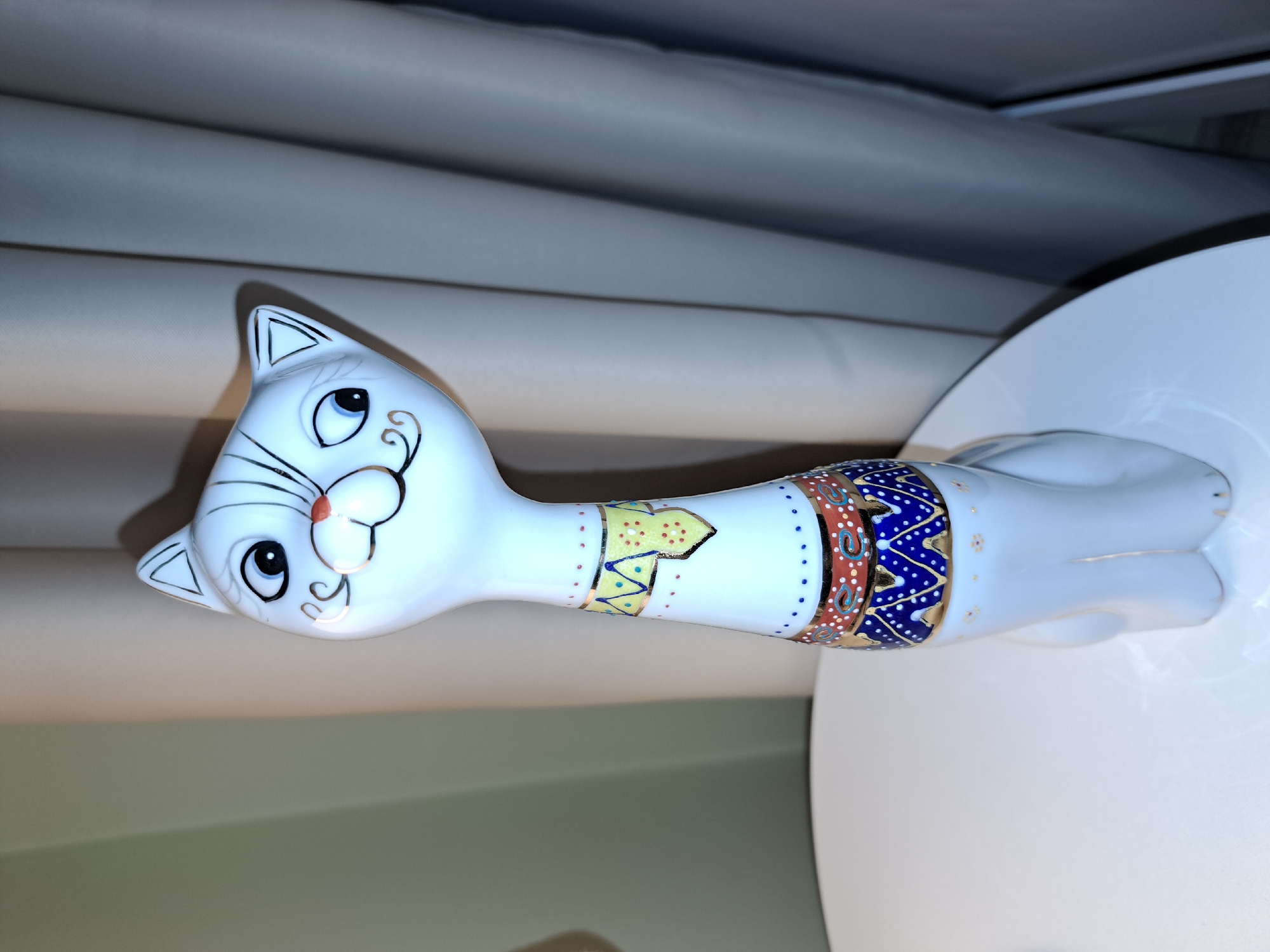 A white ceramic cat figurine with black and gold accents and a colorful patterned collar stands on a white table in front of beige curtains. The cat has large blue eyes, a red nose, and black lines drawn around its eyes and whiskers. Its collar is decorated with yellow, blue, and red patterns. The background consists of draped curtains, suggesting an indoor setting. The image does not provide any information about its location in Irkutsk, Russia.