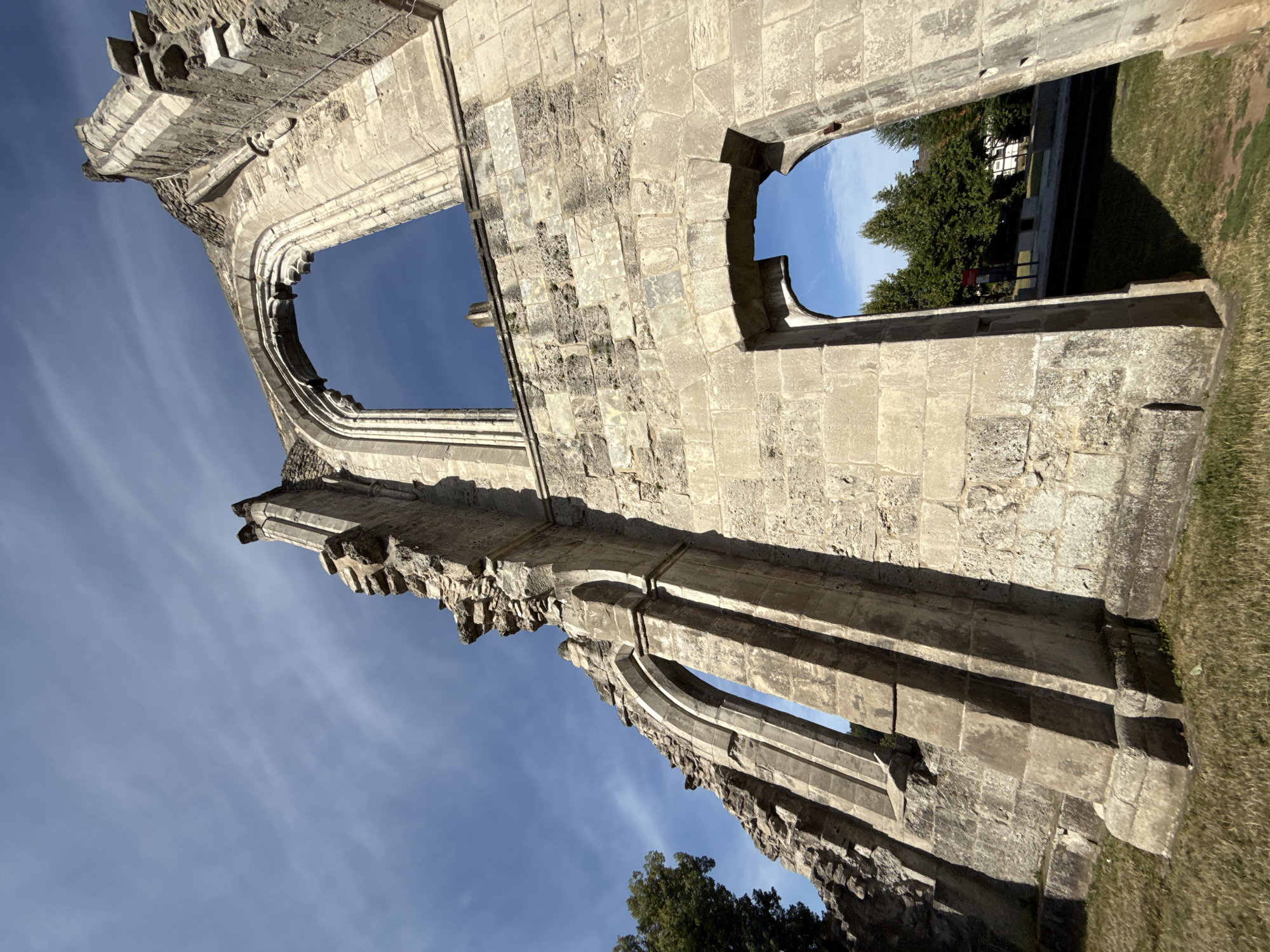 The image captures a low-angle perspective of the ruins of the Walkenried Abbey in Walkenried, Germany. The ancient stone structure, characterized by crumbling walls and arched windows, stands against a bright blue sky with wispy white clouds. Sunlight casts shadows on the weathered stone, highlighting the textures and the architectural details of the Gothic style. The foreground shows dry grass and patches of dirt. Through the openings in the ruins, glimpses of green trees, a road, and buildings can be seen in the background, suggesting the ruins are situated near a developed area. There are no people or animals visible in the image.