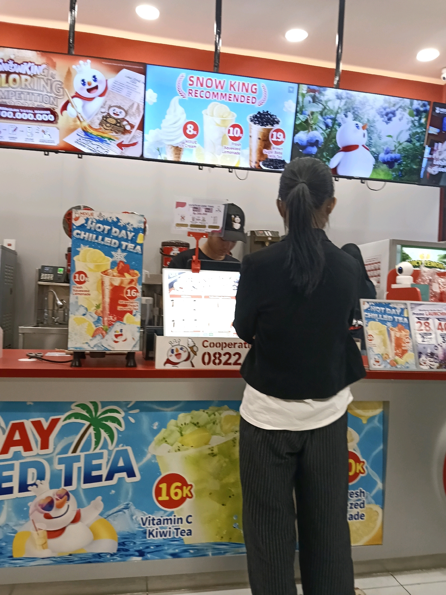 A young woman with dark hair pulled back in a ponytail stands facing away from the camera, wearing a black blazer over a white shirt and striped black pants. She is at a counter in what appears to be a food or beverage establishment. In the background, several digital screens display advertisements. One screen shows a cartoon snowman wearing a red scarf and sunglasses, floating in water with a palm tree and the text "DAY ED TEA". Another screen shows ice cream cones and drinks with prices. A sign on the counter displays "HOT DAY CHILLED TEA" with images of drinks. A man wearing a black baseball cap is partially visible behind the counter. The overall environment suggests a commercial setting, possibly a cafe or a quick-service restaurant, with bright lighting and colorful displays.