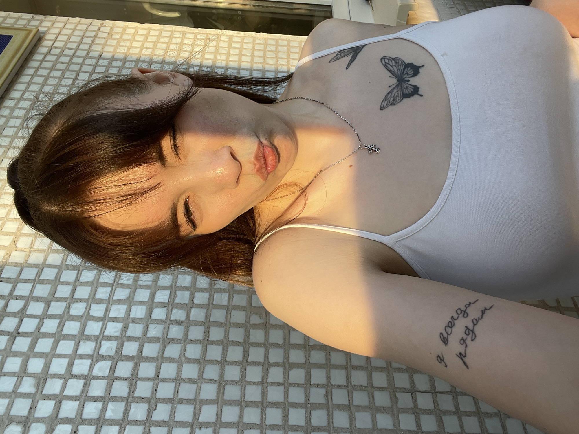 A young woman with brown hair and a white tank top is lying on a white tiled surface. She has tattoos on her chest (butterflies) and forearm (script). A silver necklace with a cross pendant is visible. The background appears to be a sunlit outdoor setting, likely a balcony or patio, featuring a glazed-window. Specific location data is unavailable.