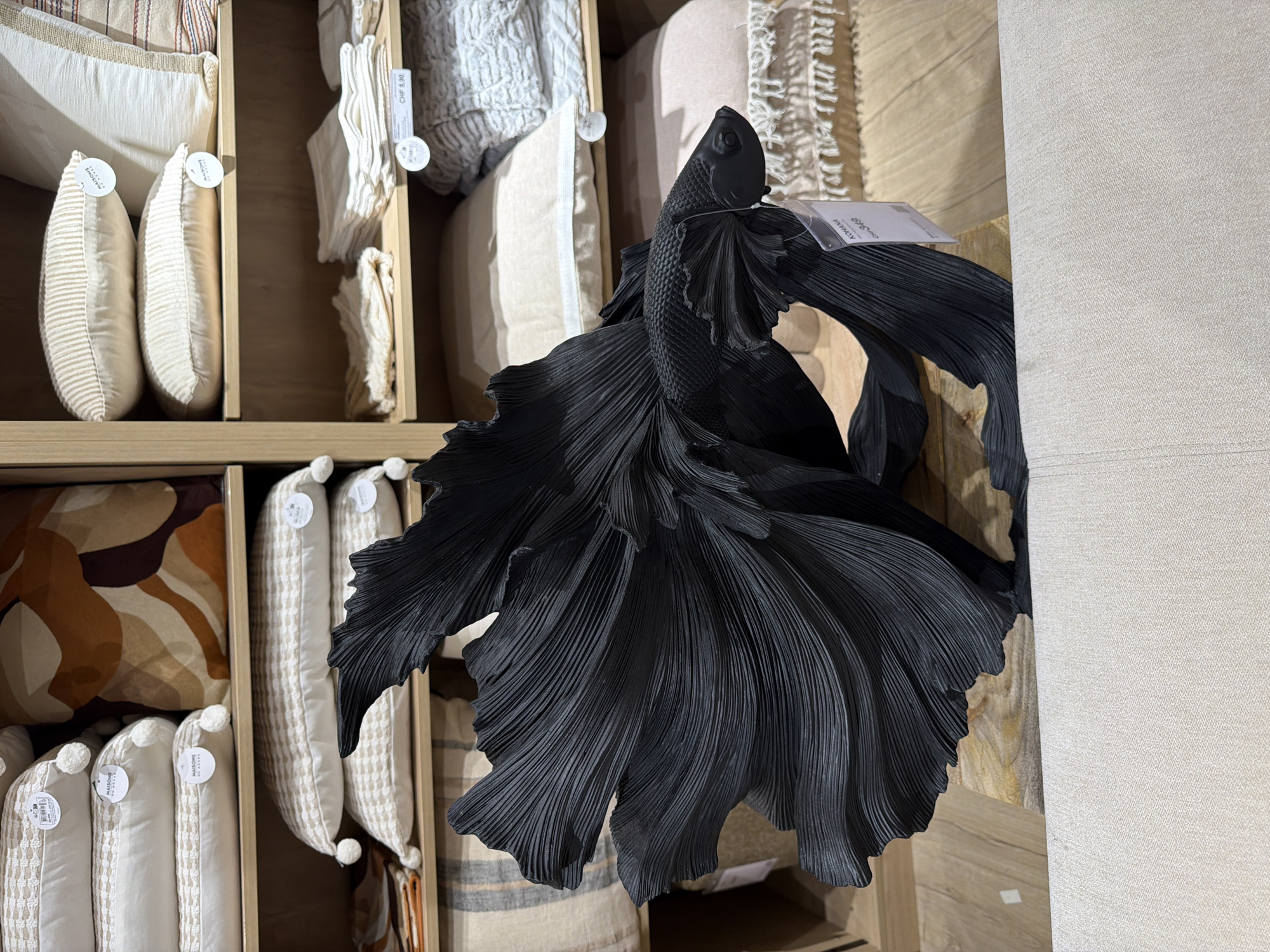 The image shows a black fish sculpture placed on a wooden surface in front of shelves filled with pillows and folded textiles. The sculpture is highly detailed, with textured fins that mimic flowing fabric. The shelves behind it contain various cushions and fabric items in neutral tones. The overall setting appears to be a retail store display.