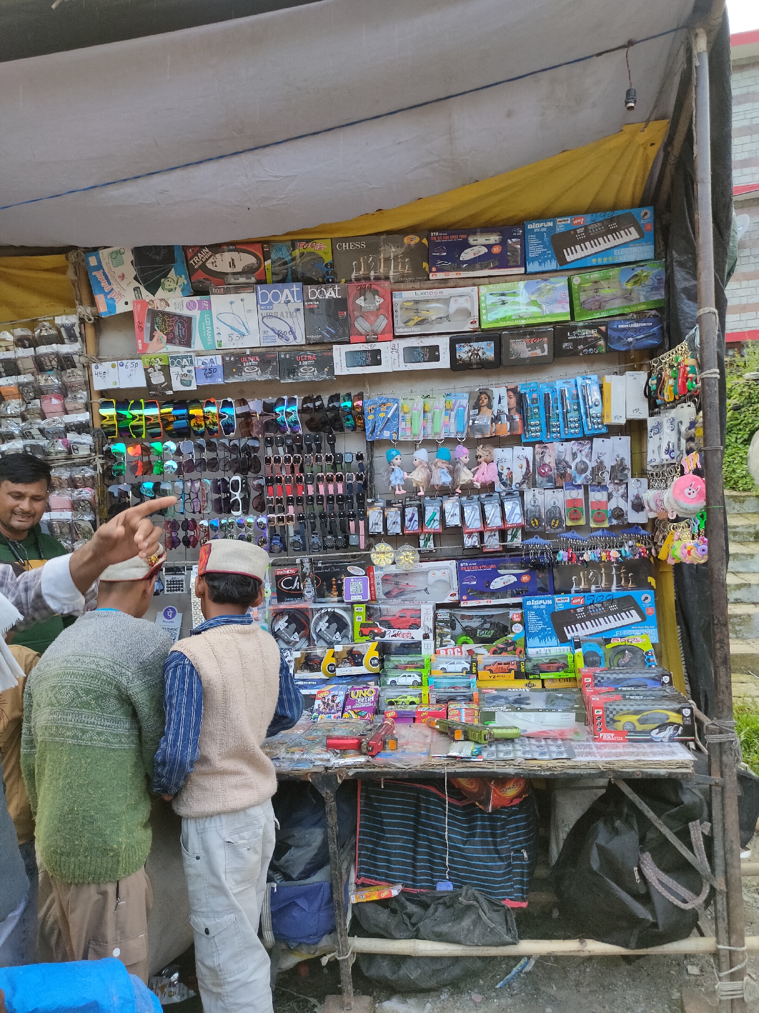 This is a full-shot, eye-level photograph of a street vendor's stall in Burwa, India. The stall is covered by a yellow awning and appears to be selling a variety of small electronics, toys, and accessories. A vendor, a man with short dark hair wearing a green shirt, is standing on the left, gesturing with his left hand. Two young boys, one in a green and white sweater and the other in a beige vest over a striped shirt, are standing in front of the stall looking at the merchandise. The stall is packed with items such as toy cars, drones, headphones, sunglasses, keychains, and a toy keyboard. The background shows a brick building and some greenery. The overall scene depicts a bustling marketplace with goods displayed for sale.