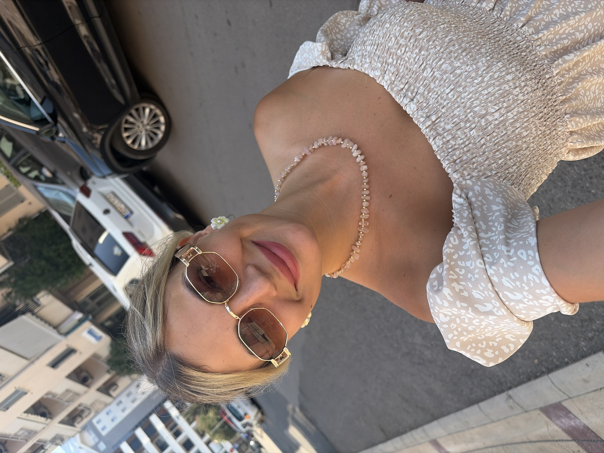 A woman with blonde hair and sunglasses is taking a selfie outdoors in Budva, Montenegro. She is wearing a light-colored, off-the-shoulder dress with a smocked bodice and a speckled pattern, and a pink beaded necklace. She is smiling at the camera. In the background, there are buildings and parked cars on a paved street.