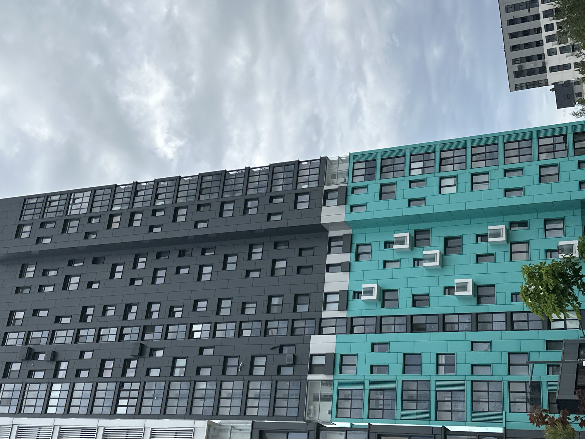The image depicts a multi-story building located in Бараўлянскі сельскі Савет, Belarus. The building's exterior features two distinct sections: one is a dark grey and the other is a vibrant turquoise color. Both sections are adorned with numerous windows arranged in a grid-like pattern. On the turquoise section, some of the windows are accompanied by small, white protruding structures. The sky above is overcast with grey clouds. Another building, appearing to be white and grey, can be seen in the upper right corner of the image. Some tree foliage is visible along the right edge of the building.