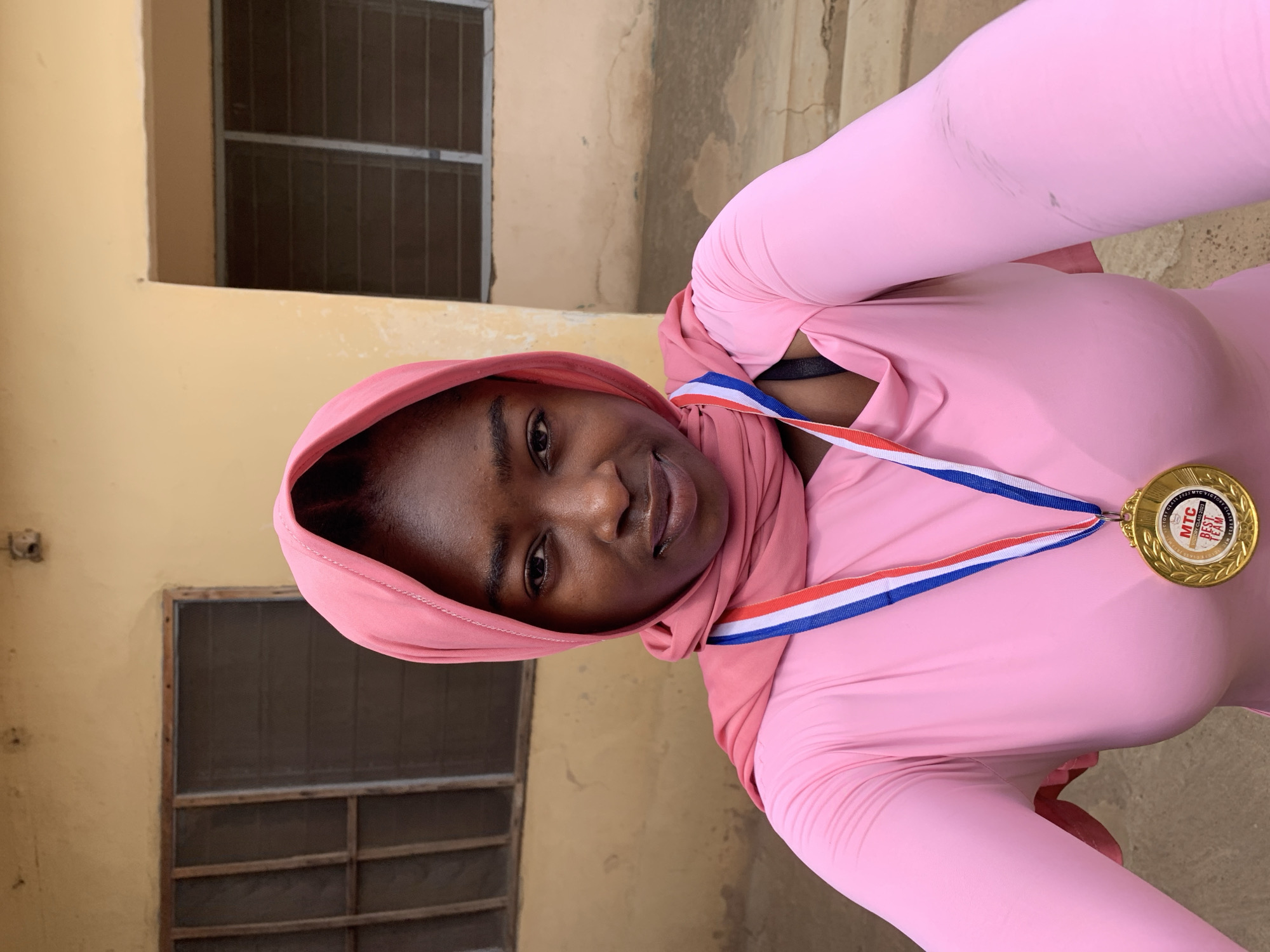 A young, dark-skinned woman wearing a pink headscarf and a pink long-sleeved outfit is the subject of a selfie. She is smiling and looking at the camera. A gold medal with a blue, white, and red ribbon hangs around her neck.
The background shows a beige building exterior with barred windows. The location is Rafin Par, Nigeria, based on the medal.